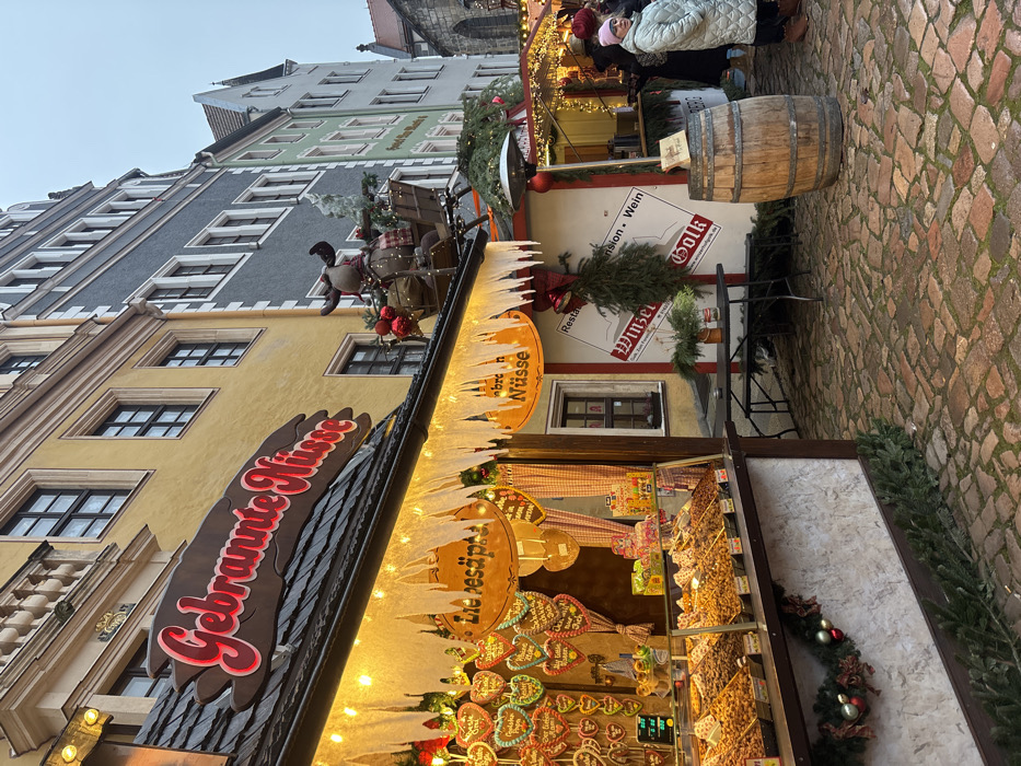 The image displays an outdoor Christmas market scene in Meissen, Germany, with a cobblestone ground under an overcast sky.
In the foreground and midground, a market stall prominently features the illuminated sign "Gebrannte Nüsse" (roasted nuts) in red script with a white border on a dark brown backdrop. The stall's awning is adorned with white, icicle-shaped cutouts and string lights. Its display shelves are laden with various roasted nuts and candies. Gingerbread hearts, some labeled "Liebesäpfel" and others with German messages, hang from the interior. A festive garland with red spherical ornaments drapes the lower front of the stall. A plush reindeer figure, dressed in a plaid scarf and pulling a small sleigh containing red ornaments and greenery, is mounted on the roof above the right side of the "Gebrannte Nüsse" stall.
To the right, a black folding table with benches is situated on the cobblestones. Adjacent is a second, partially visible market stall, displaying a sign reading "Winzer...sion - Wein," decorated with evergreen branches and red bells. A weathered wooden barrel stands directly in front of this stall.
One female individual is visible in the midground to the right, wearing a light-colored, possibly mint green, long puffer coat and a pink knit beanie. Her head is turned slightly to the left. Blurred figures are discernible in the deeper background.
The background features a row of multi-story buildings: a yellow-facade building directly behind the "Gebrannte Nüsse" stall, followed by a gray building, then lighter green and cream-colored structures, all with visible windows. A dark, gothic-style architectural structure with a pointed roof is partially visible in the far background on the right.