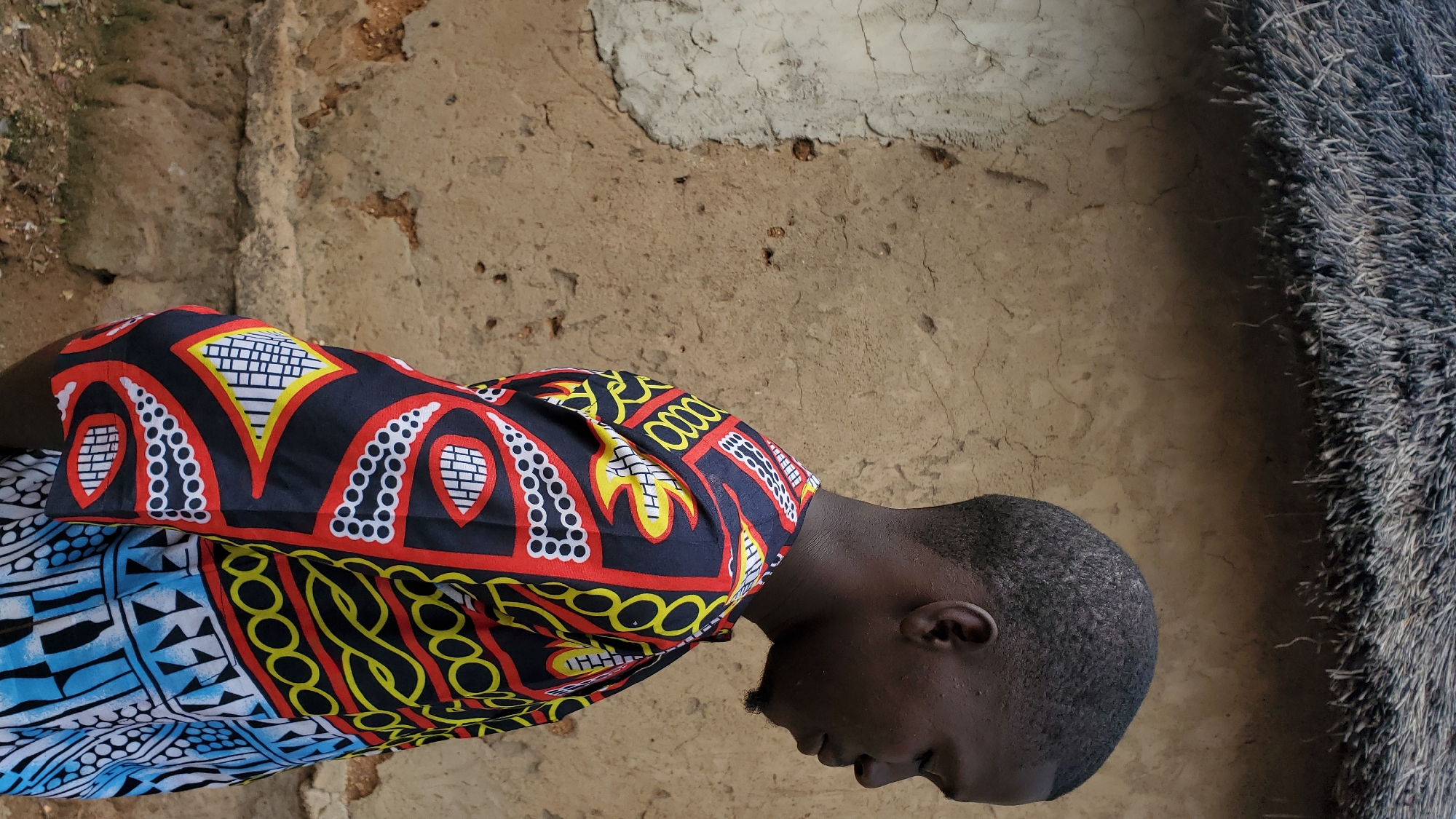 A young Black man is pictured in a medium close-up, facing right with his eyes closed and head slightly bowed. He wears a colorful garment with a black base and a vibrant pattern of red, yellow, and white geometric and abstract shapes. The man has short, dark hair and a faint beard. He stands against a textured, earth-toned wall that appears to be made of mud or stucco. Above the wall, a thatched roof made of dry reeds is visible. The setting suggests an outdoor environment, possibly in a rural area or a less developed part of a city, consistent with the visual cues of traditional building materials and natural roofing found in places like Jalingo, Nigeria. The lighting is soft, indicating an overcast day or the shaded side of a building.