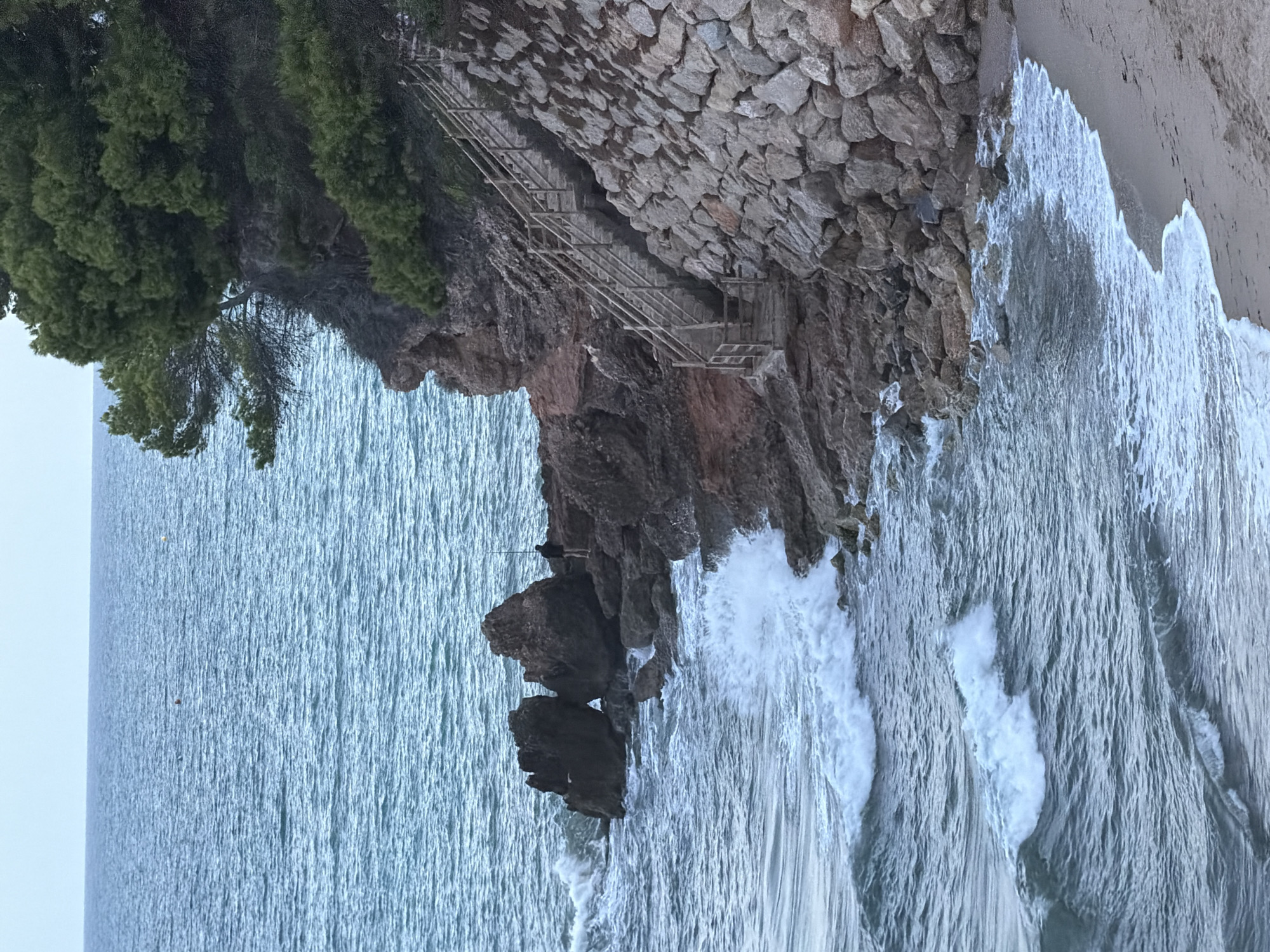 The image depicts a rugged coastal scene near Mont-roig del Camp, Spain. In the foreground, a sandy beach is met by the gentle lapping of waves. To the left, the sea is a deep blue, with white foam cresting on incoming waves. Jagged, dark rock formations jut out from the water, some partially submerged. On the rocky coastline, a lone individual is visible, standing on a ledge and appearing to be fishing with a rod. To the right, a steep, rocky incline has been reinforced with a wall of large stones. A wooden staircase with railings ascends this incline, leading up to a dense growth of pine trees and other vegetation crowning the cliff. The sky is overcast, casting a diffused light over the scene.