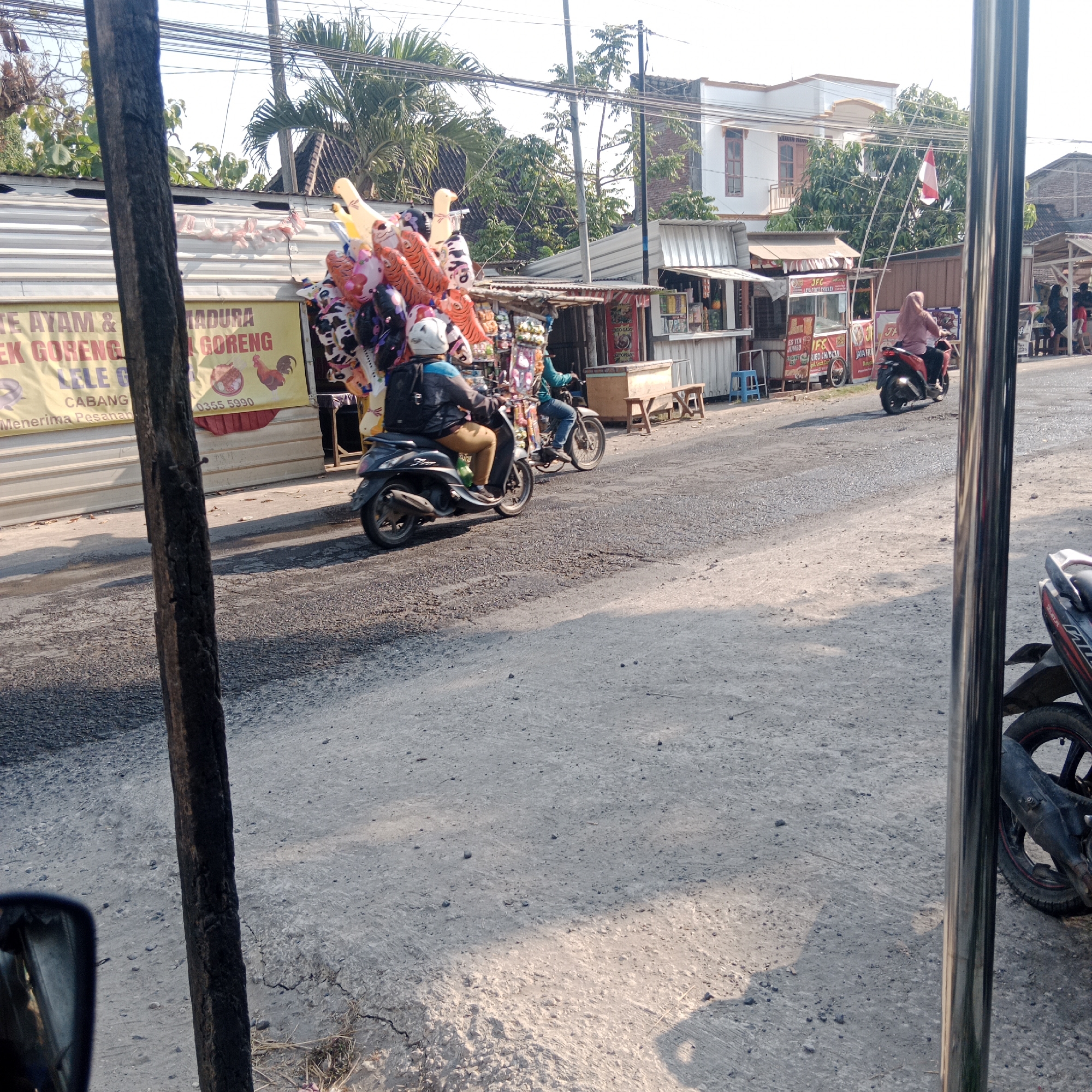 This is a street scene in Tegalrejo, Indonesia. A wooden pole is in the foreground on the left. A man wearing a white helmet, blue jacket, and brown pants is riding a dark-colored scooter on the asphalt road. He is carrying a large bunch of colorful balloons, including tiger-striped, cow-spotted, and yellow duck-shaped ones. Behind him, another person on a scooter is visible. To the right of the road, there are several small food stalls with white corrugated metal roofs. One stall has a yellow banner advertising "AYAM & BEBEK GORENG" (fried chicken and duck) and "LELE GORENG" (fried catfish), with phone number "0355 5990". Another stall displays a flag. Further down the road, a woman wearing a pink hijab is riding a red scooter. In the background, there are trees and multi-story buildings. The ground is paved with asphalt and gravel.