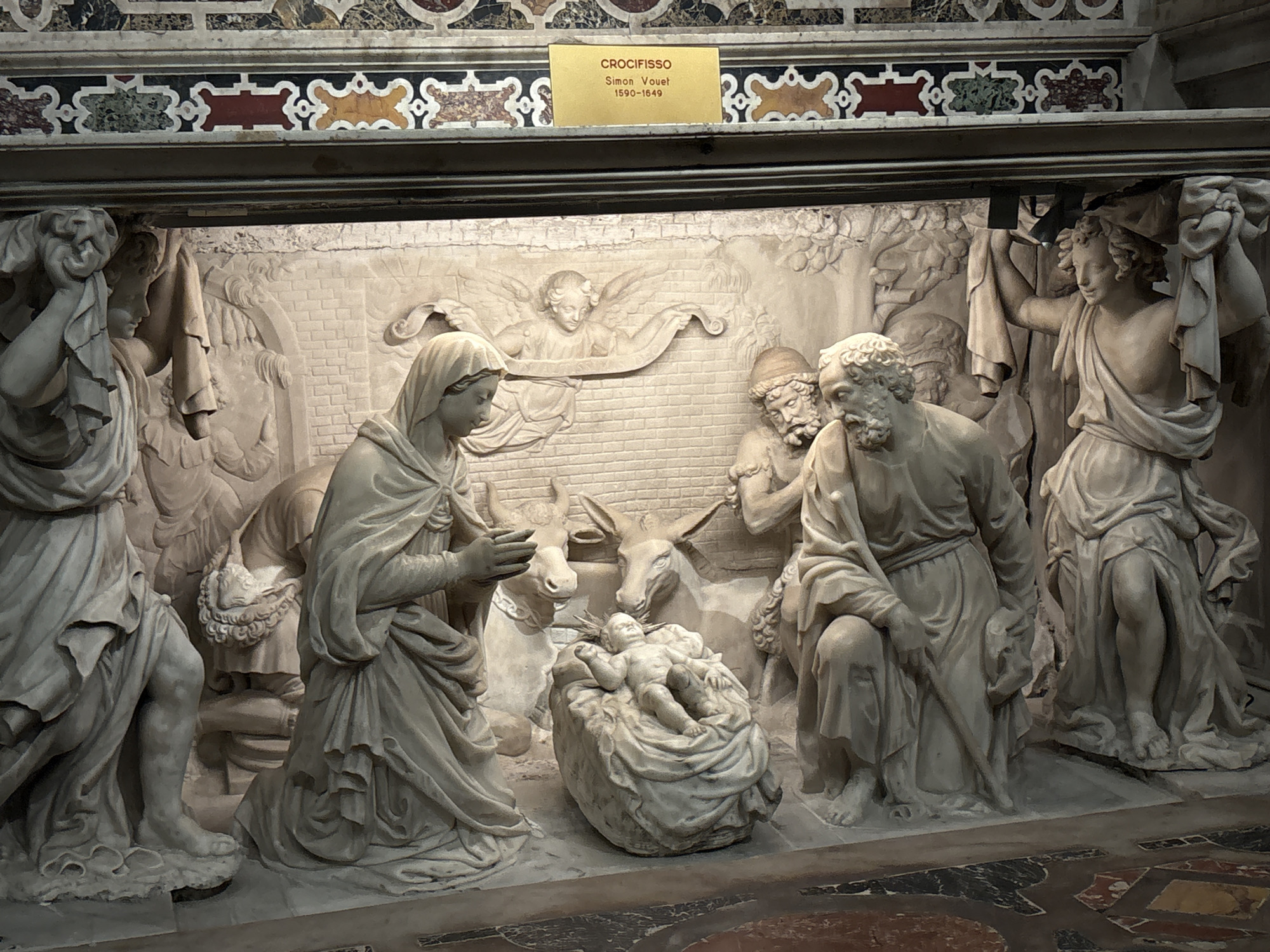The image depicts a marble bas-relief of the Nativity scene. The scene includes Mary kneeling and looking at the baby Jesus lying in a manger. Joseph kneels beside them. Other figures surround them, including angels and shepherds with donkeys. The bas-relief is under a decorative architectural border of colorful marble. A small sign above the sculpture reads "CROCIFISSO Simon Vouet 1590-1649." This is located in Genoa, Italy.