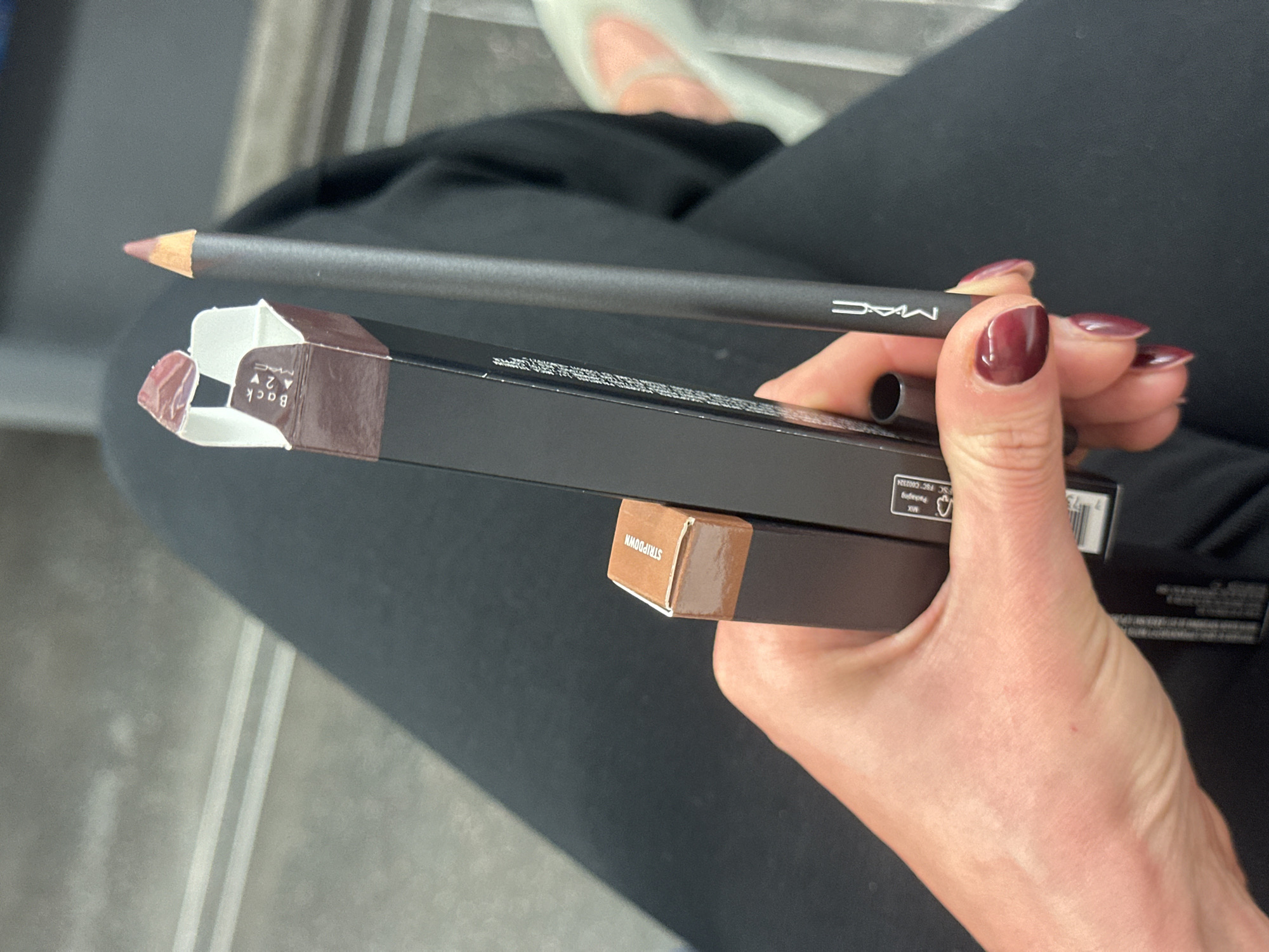 A slightly overhead, close-up shot captures a person's hand holding makeup products. The hand, adorned with deep burgundy nail polish, firmly grasps a dark grey MAC lip pencil with a reddish-brown tip. Beneath the pencil, the hand holds two black rectangular MAC product boxes. One box is partially open, revealing an internal flap that reads "BACK" (upside down) and "A2V." The side of the second box displays the product name "STRIPDOWN." Other visible text includes the "MAC" logo on the pencil and boxes, and partially obscured details like "MIX" and "FSC C002324" along with a barcode on one of the boxes.
The person is seated, with their black-clad legs and a foot in a light-colored shoe visible in the background. The textured grey floor beneath features distinct parallel white lines, suggesting an indoor public setting like a train carriage, bus, or a waiting area. The lighting is consistent with an indoor environment, either during the day or under artificial illumination. The overall scene depicts the act of holding and examining new beauty products. The context places this scene in London, United Kingdom.