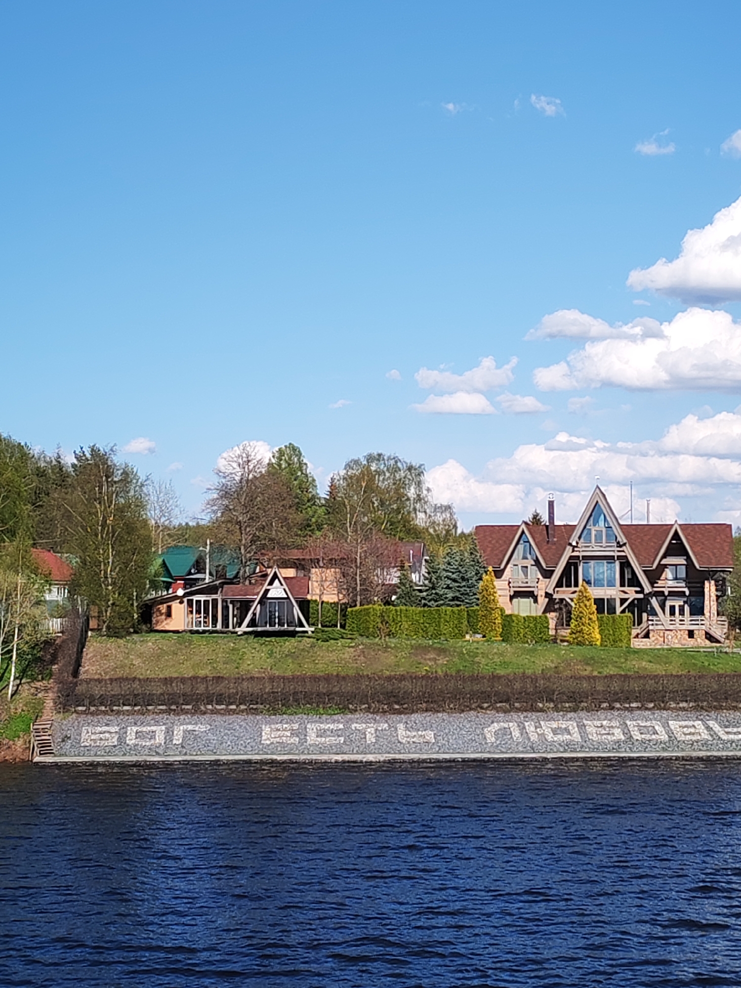 The image shows a waterfront view with several buildings on a grassy rise. The primary structure is a large, modern house with a prominent triangular roofline. To the left are other houses and trees. A low wall runs along the waterfront, bearing a mosaic inscription written in Cyrillic. The water is dark and reflects the sky. The sky is blue with scattered white clouds. The scene is located in Tarchevo, Russia based on the text on the wall.