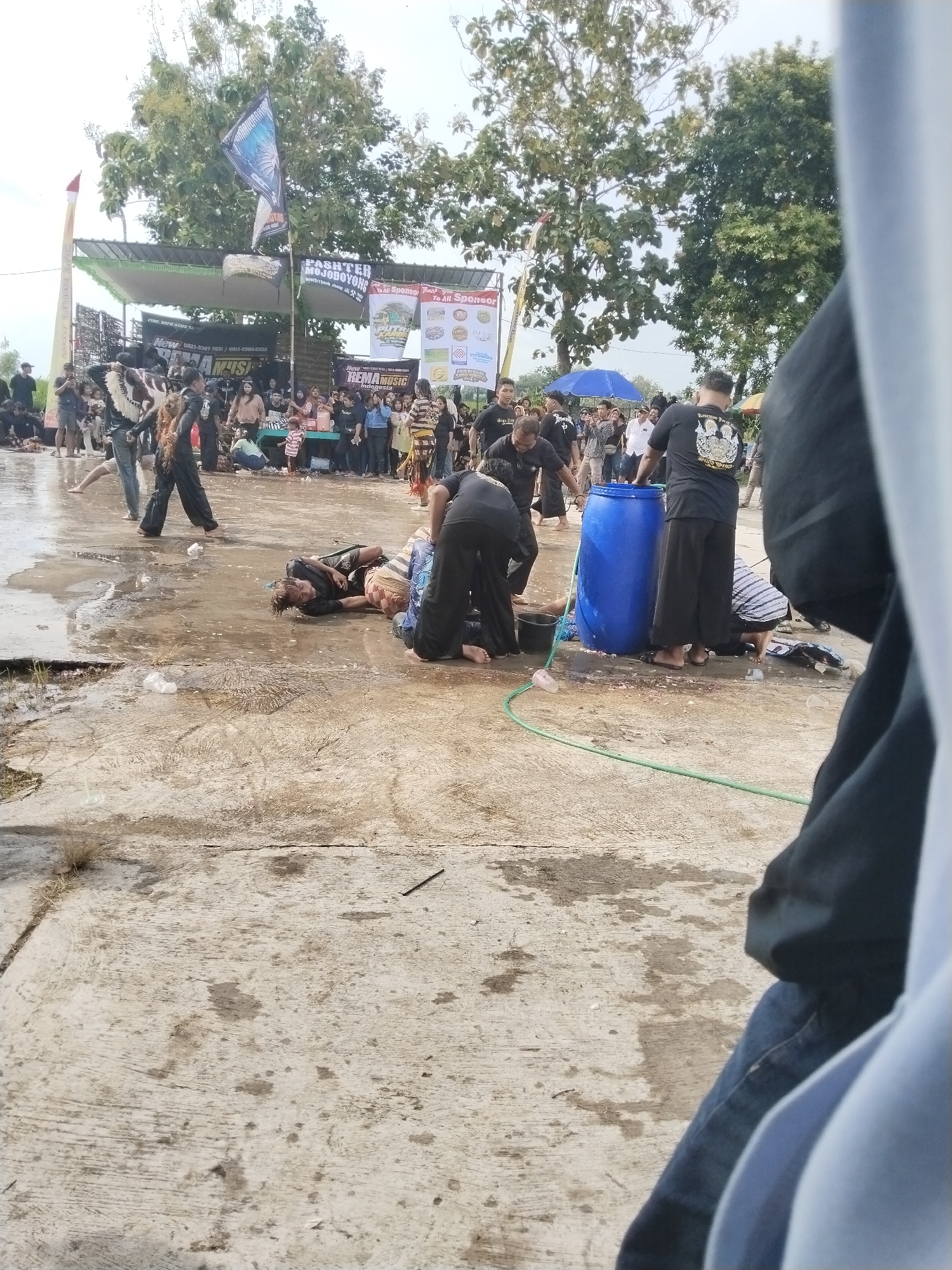 The image depicts an outdoor event, possibly a cultural performance or competition, taking place in what appears to be Mojodoyong, Indonesia, given the visible signage. A crowd is gathered around a stage in the background, and a blue barrel and green hose are present in the foreground. Several people, dressed in black, are tending to individuals lying on the ground in a wet area, suggesting they might be injured or part of a performance. A banner with text, including "PASHTER MOJODOYONG" and "REMA MUSIC Indonesia," is visible on the stage. The ground is concrete and appears wet.