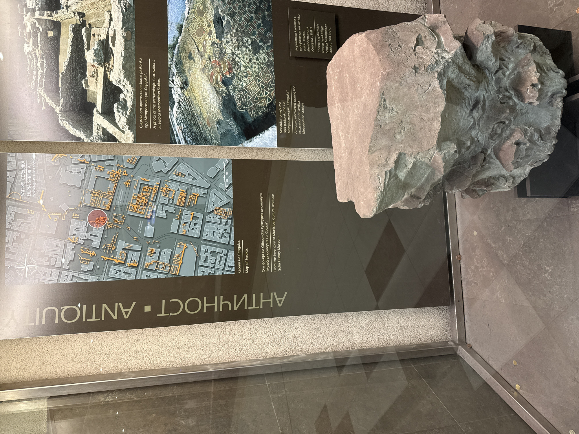 The image depicts a museum exhibit showcasing artifacts from the ancient city of Serdica, now Sofia, Bulgaria. The exhibit includes a sign in Bulgarian and English that reads "ANTIQUITY" and provides details about the artifacts displayed.
On the sign, there is a photo of archaeological excavations at the Serdica Metropolitan Station, and another photo of a mosaic uncovered during the excavations. The sign also includes a map of Serdica, with the location of the city's ancient ruins marked in orange. The map's title is "Map of Serdica."
To the right of the sign, a stone object that appears to be a Corinthian capital with female head effigy (2nd-3rd c.).
The exhibit is housed in a glass display case, and the overall environment is museum-like.