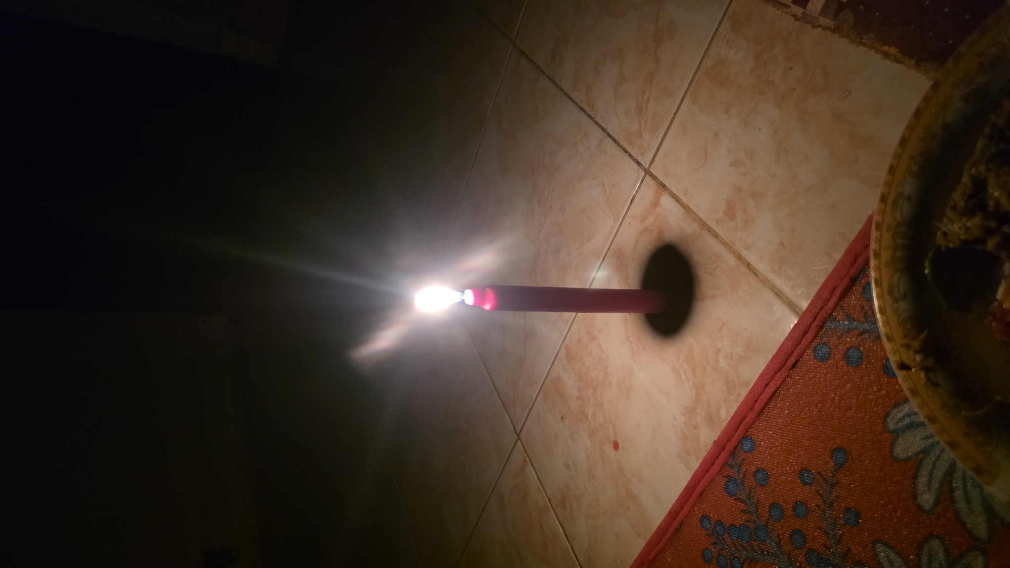A lit red candle casts a bright glow and a distinct shadow on a tiled floor. A mat with an orange background and a blue floral design is partially visible in the lower right corner of the frame, next to a plate. The left side of the image is shrouded in darkness. This image was taken in Lhokseumawe, Indonesia.