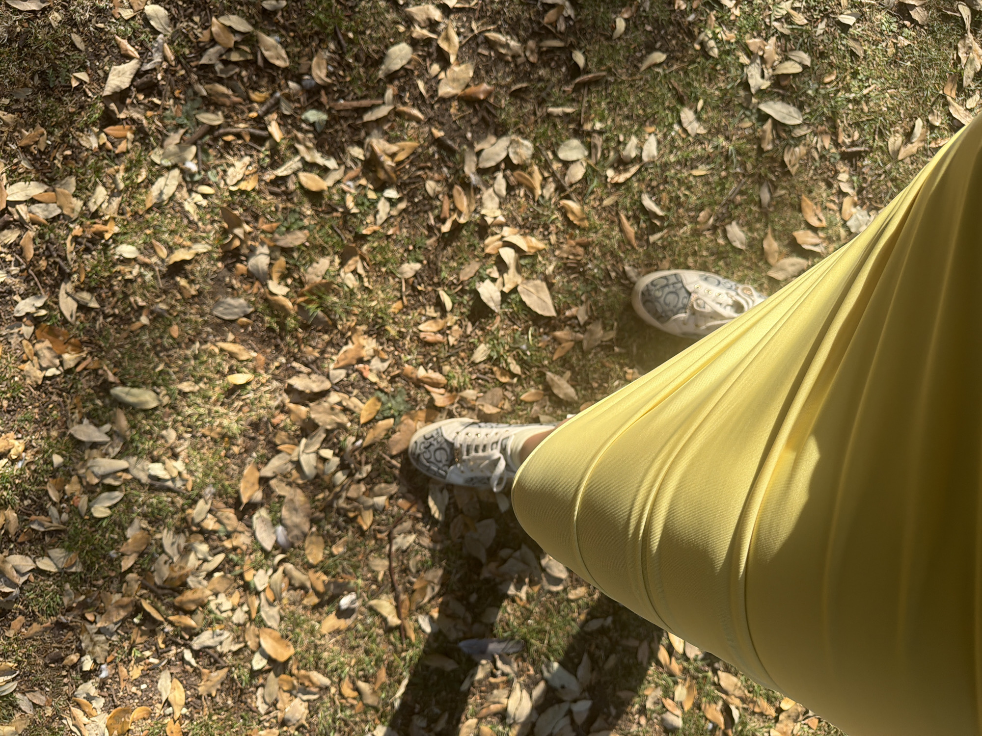 The image presents a view from above, looking down at a section of ground covered in fallen leaves mixed with green grass. Two feet wearing sneakers are visible amidst the foliage. A large expanse of yellow fabric dominates the right side of the frame, likely a portion of a garment such as a skirt or dress. The scene is bathed in natural light. No other people or objects are clearly discernible. There is no information in this image to suggest its location is London, United Kingdom.