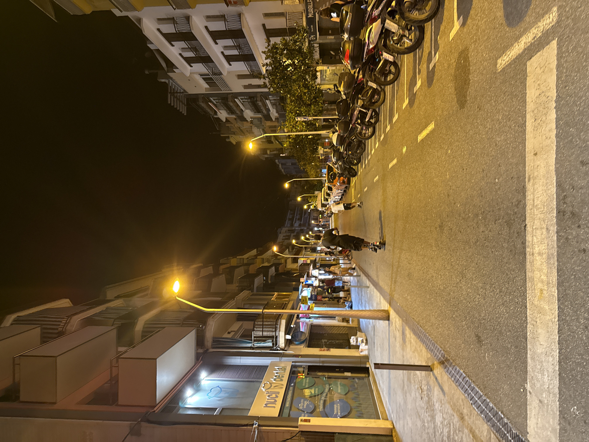 An outdoor, nighttime street scene in Blanes, Spain features buildings lining both sides of a paved road. The buildings appear to be multi-story with balconies and illuminated windows. A streetlamp with a bright yellow light is visible in the foreground, casting a glow on the street. A person is riding an electric scooter down the middle of the street. Parked motorcycles are lined up along the right side of the road. Pedestrians can be seen on the sidewalk on the left side of the street, and a business with illuminated signage is also present. The street has white markings indicating parking spaces and a pedestrian crossing. Trees are planted along the sidewalk.