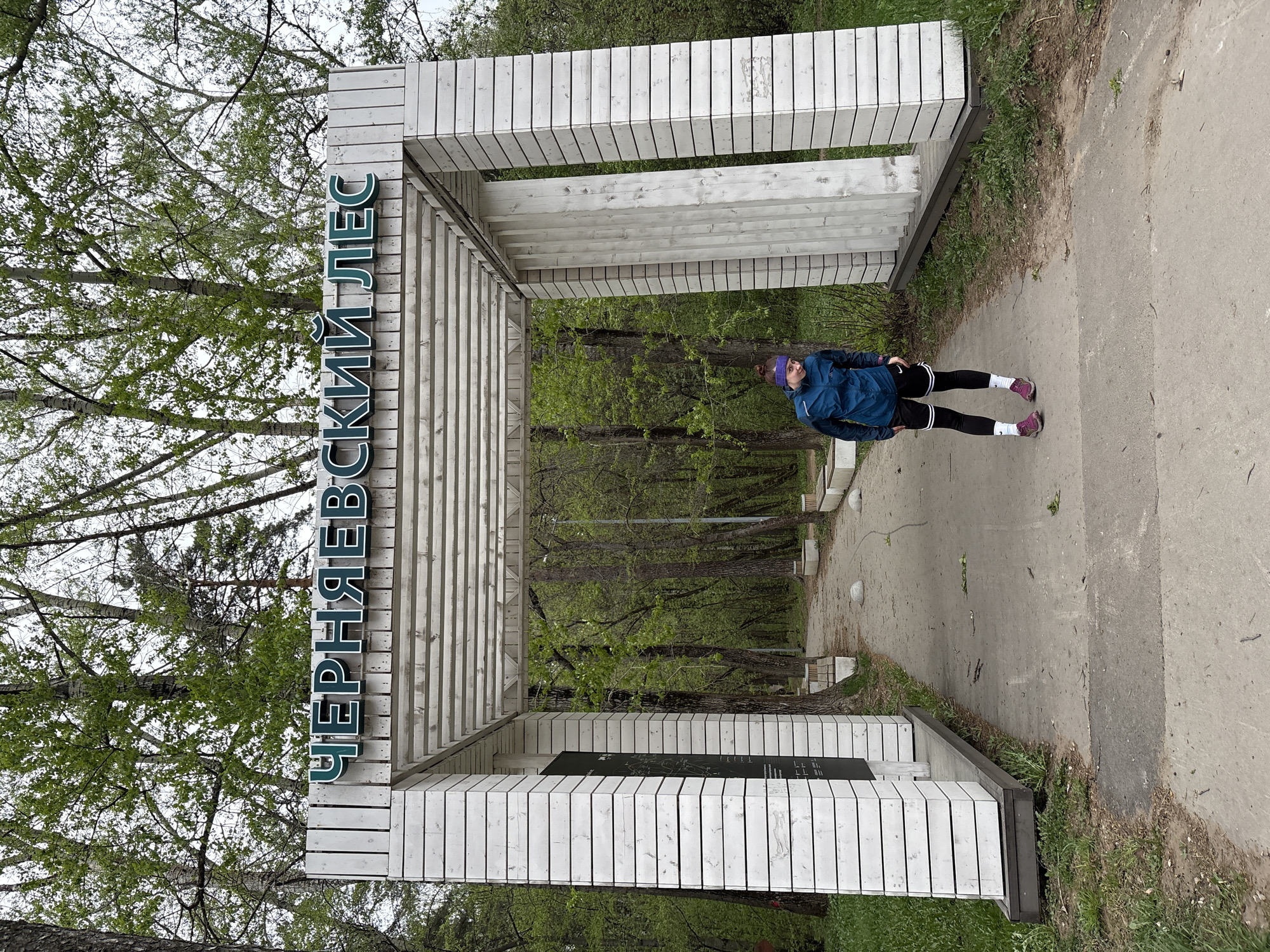 A medium shot presents a woman standing within the entrance structure of the Chernyaevsky Forest in Perm, Russia. The structure is constructed from white wooden planks forming a pergola-like gateway. The name "ЧЕРНЯЕВСКИЙ ЛЕС" (Chernyaevsky Forest) is written vertically in green letters. The woman is wearing a blue jacket, black leggings with white socks, and a purple hat. She is standing on a concrete path that extends into the forest. The forest appears lush with greenery, indicating the presence of trees. The sky above is overcast.