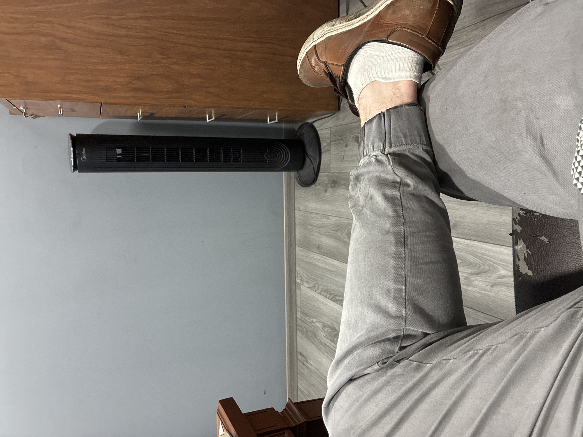 A medium-angle shot depicts a person sitting, with their right leg visible. The individual is wearing a white sock and a brown leather shoe. They are dressed in gray pants with elastic cuffs. A black tower fan is positioned on the floor near the leg. The environment includes a light gray wall and a wooden surface above the fan. The location is possibly Cuautitlán Izcalli, Mexico, but this cannot be confirmed from the image.