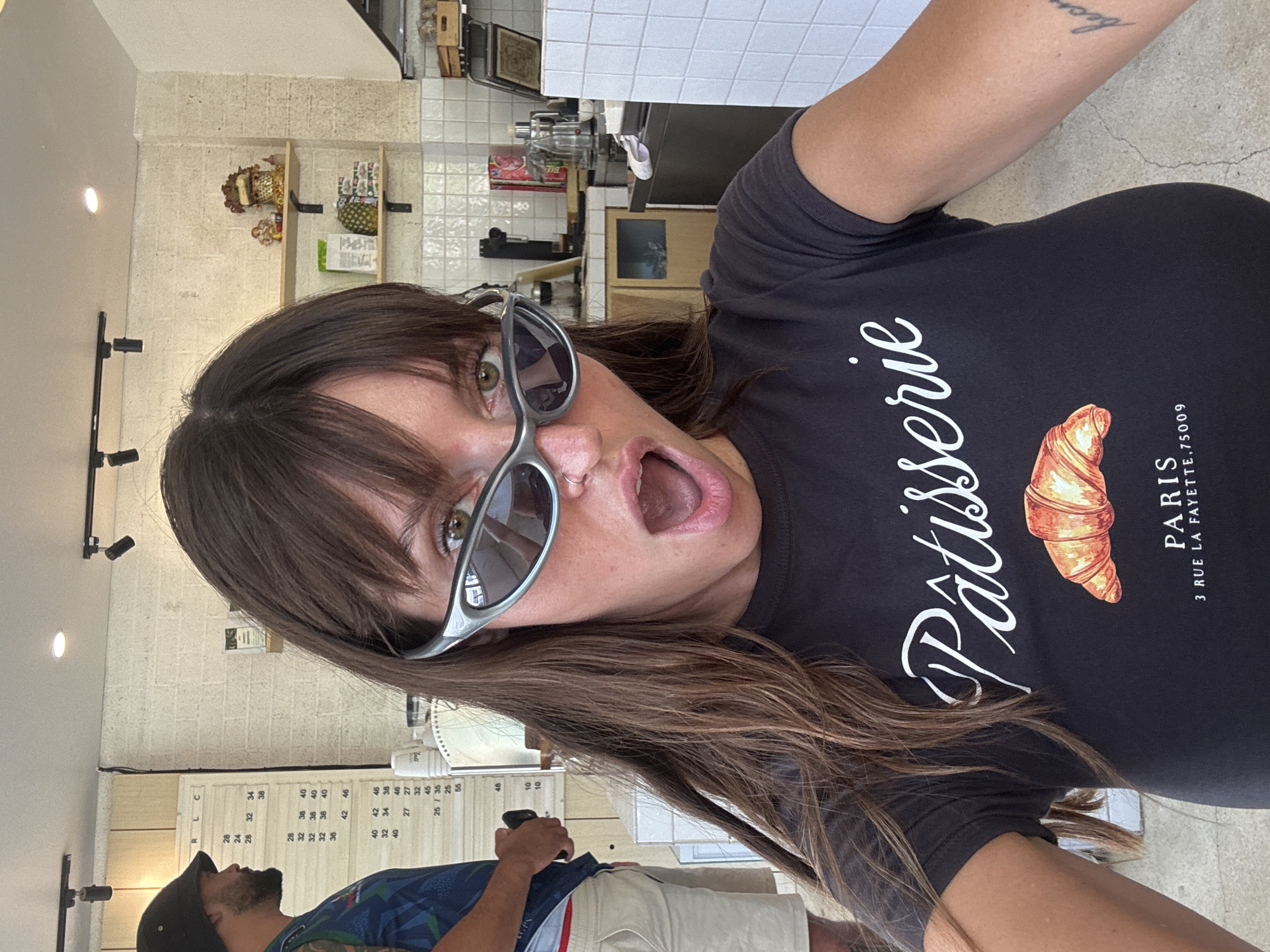 The image shows a woman with brown hair and bangs, wearing sunglasses and a black t-shirt with "Pâtisserie PARIS 3 RUE LA FAYETTE,75009" printed on it, making a surprised face. The background appears to be a pastry shop or cafe, with a menu board visible to the left and shelves with items behind the counter. A man wearing a hat and colorful shirt is visible partially in the lower left corner. The photo is taken indoors, with fluorescent lighting. The specific location in North Kuta, Indonesia is not evident from the image.