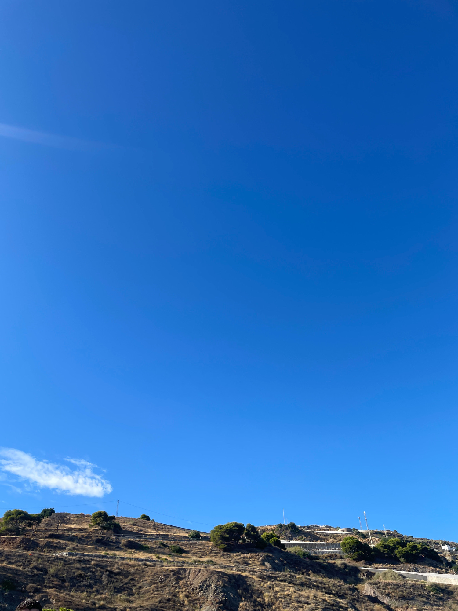 The image shows a clear blue sky over a dry, hilly landscape. The terrain is covered in sparse, brown vegetation, with scattered trees dotting the slopes. On the crest of the hill, several white structures are visible, possibly buildings or infrastructure. A power line stretches across the hillside. In the lower left corner of the frame, a small portion of a person's head and shoulder is visible, suggesting they are in the foreground. The scene appears to be an outdoor view in a dry, sunny environment. The location is Lújar, Spain.
