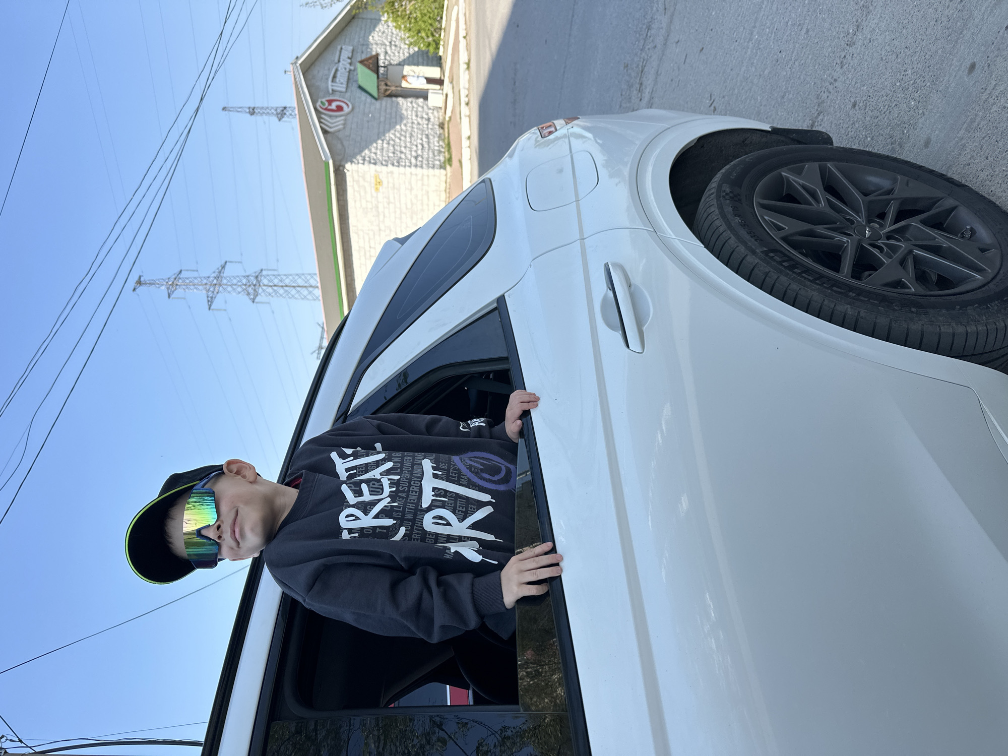 A boy in a black cap and reflective sunglasses leans out of the open window of a white car. He wears a black sweatshirt with the word "CREATE" printed on it. The car is parked near a building with a sign that reads "Primeram" in Cyrillic. Overhead power lines and pylons are visible in the background. The location is in the city of Chaikovsky, Russia.