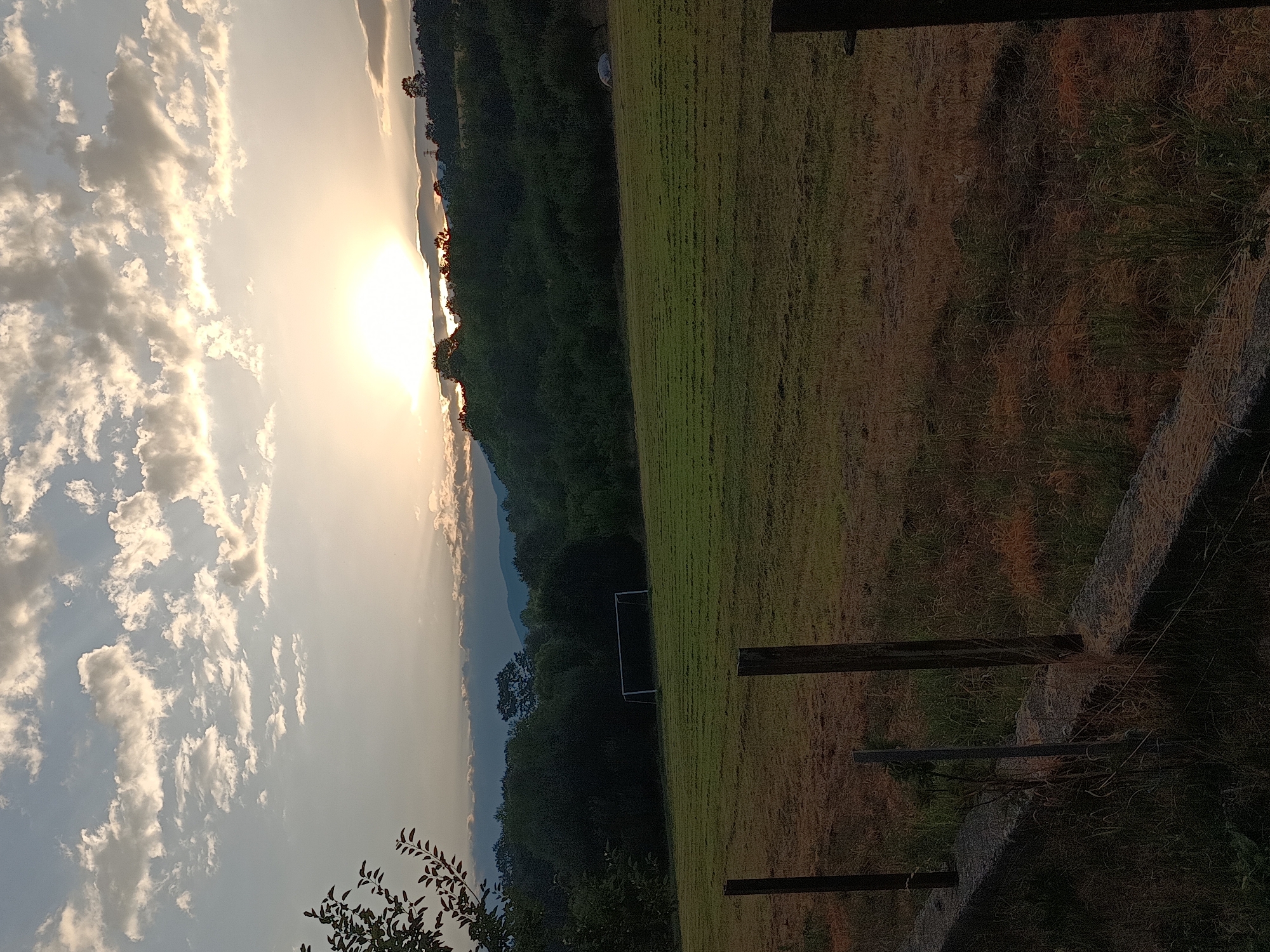The image depicts a pastoral scene at dusk, likely in Kralev Dol, Bulgaria, given the context provided. The foreground features a low concrete wall with wooden posts extending upwards, bordering a field. The field itself is predominantly dry, sun-colored grass with some areas of greener vegetation. In the middle ground, a soccer goal is visible on a well-maintained green field. Beyond this field, a dense line of trees forms a dark green horizon, with rolling hills and mountains in the distance. The sky is a prominent feature, with scattered, luminous clouds illuminated by the setting sun, which appears as a bright, soft orb near the center of the horizon, casting a warm glow. The overall atmosphere is serene and tranquil.