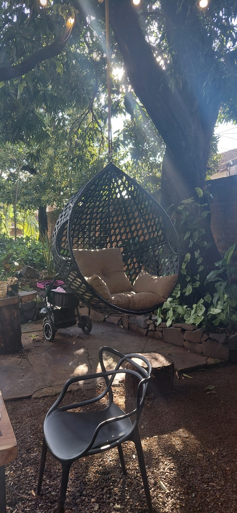 The image depicts an outdoor seating area in Encarnación, Paraguay, featuring a hanging egg-shaped chair made of dark woven material, suspended from a tree branch by a metal chain. The chair is furnished with several light-colored cushions. In the foreground, a modern black plastic chair with a distinctive curved back is positioned on a ground covered with gravel and scattered leaves. To the left of the hanging chair, a black stroller with white wheels is visible, and a wooden table top rests on a support structure. The scene is set amidst lush green foliage and a large tree trunk. Sunlight filters through the leaves, creating dappled light patterns on the ground. A brick wall is visible in the background, along with a portion of a building with a tiled roof. The overall impression is of a tranquil, shaded outdoor space.