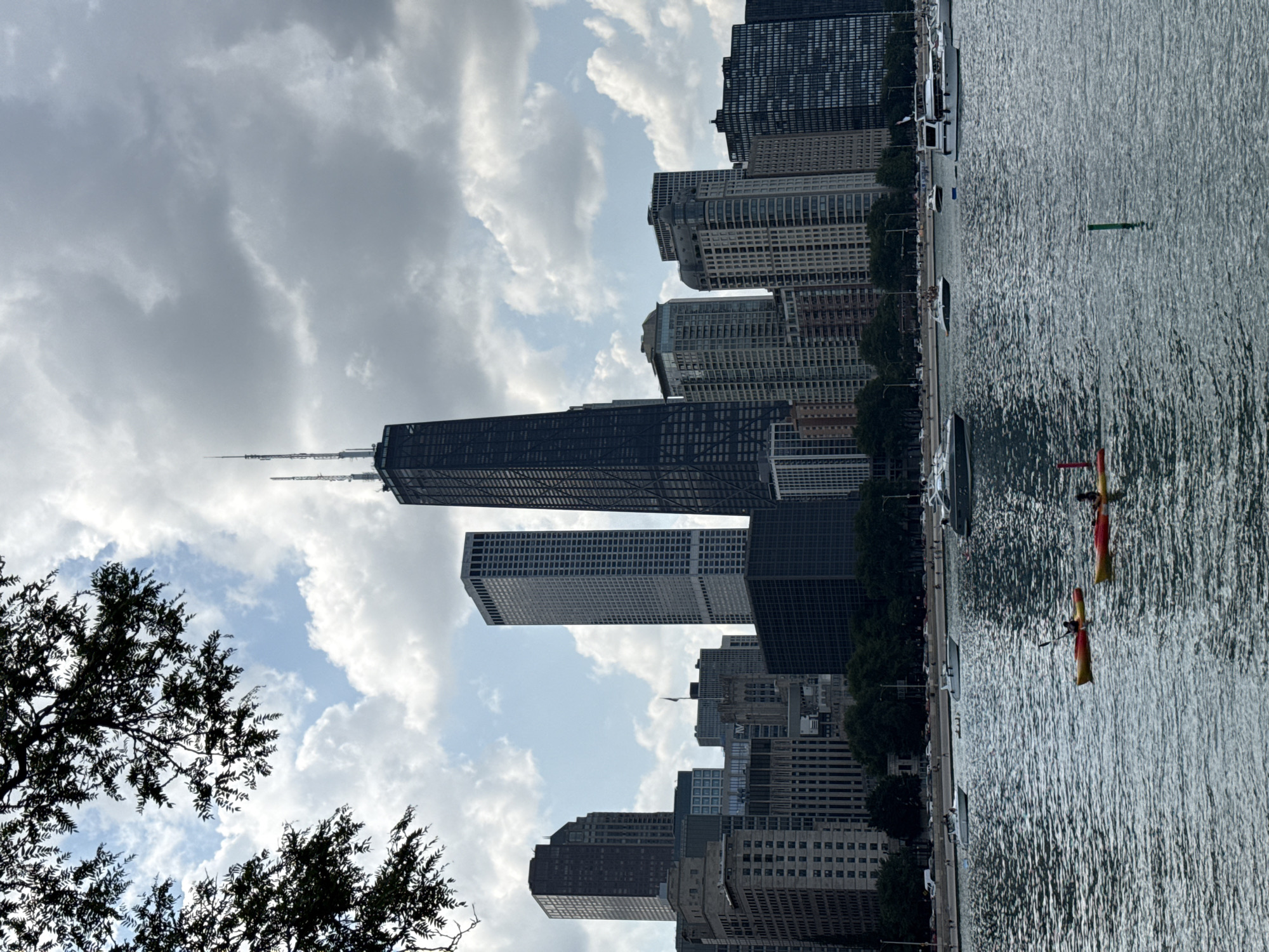 The image depicts a wide shot of the Chicago skyline from the water, likely Lake Michigan. The John Hancock Center dominates the center of the frame, a tall, dark skyscraper with a distinctive lattice exterior and two antennas at its peak. To its left and right are other skyscrapers of varying heights and architectural styles, with a mix of glass and concrete facades. In the foreground, the water is choppy with gentle waves. Several boats are scattered across the lake, including two kayaks in the middle distance, each occupied by one person. Further back, a white motorboat is visible, and in the far distance, a larger yacht is moored. Along the shoreline, a treeline separates the water from the cityscape, with a visible promenade or road behind the trees where small figures can be seen walking. The sky is partly cloudy, with dramatic cumulus clouds casting shadows on the buildings and water, suggesting a bright but potentially changeable day. A branch with dark green leaves is in the upper left corner of the image, framing the scene.