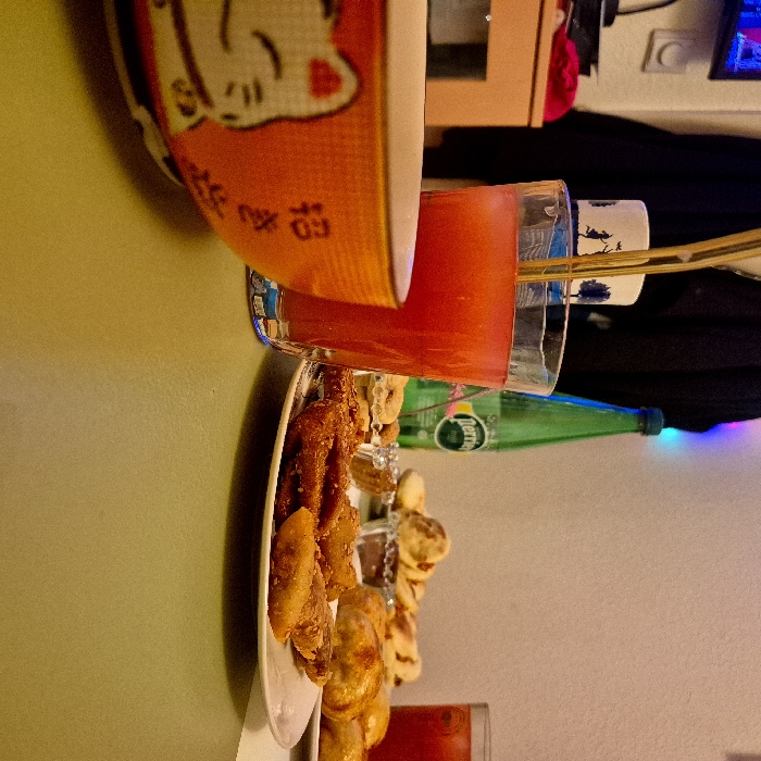 The image displays an indoor table setting in a residential or casual environment located in Strasbourg, France, captured from a low perspective. The foreground features a light olive-green table surface. On the left, an orange ceramic bowl decorated with a white Maneki-neko cat and Japanese characters is partially visible. Adjacent to it, a clear glass contains a red beverage, likely tomato juice, with two golden metallic straws and ice. To the right, a white oval plate holds multiple pieces of fried food, possibly plantains or fritters, topped with sesame seeds. Behind this plate, a green Perrier sparkling water bottle stands upright. Further back on the table, a clear glass dish or bowl contains several round pastries or dumplings and individual snacks presented in silver foil cups. Another clear glass filled with a red beverage is partially visible on the far right. The background consists of a light-colored wall, partially obscured by a dark hanging fabric. A white mug with a silhouette design is discernible behind the main glass. Faint, colored decorative lights (red, blue, green) are visible on the upper right background, indicating an evening or festive atmosphere. No individuals are present in the scene.