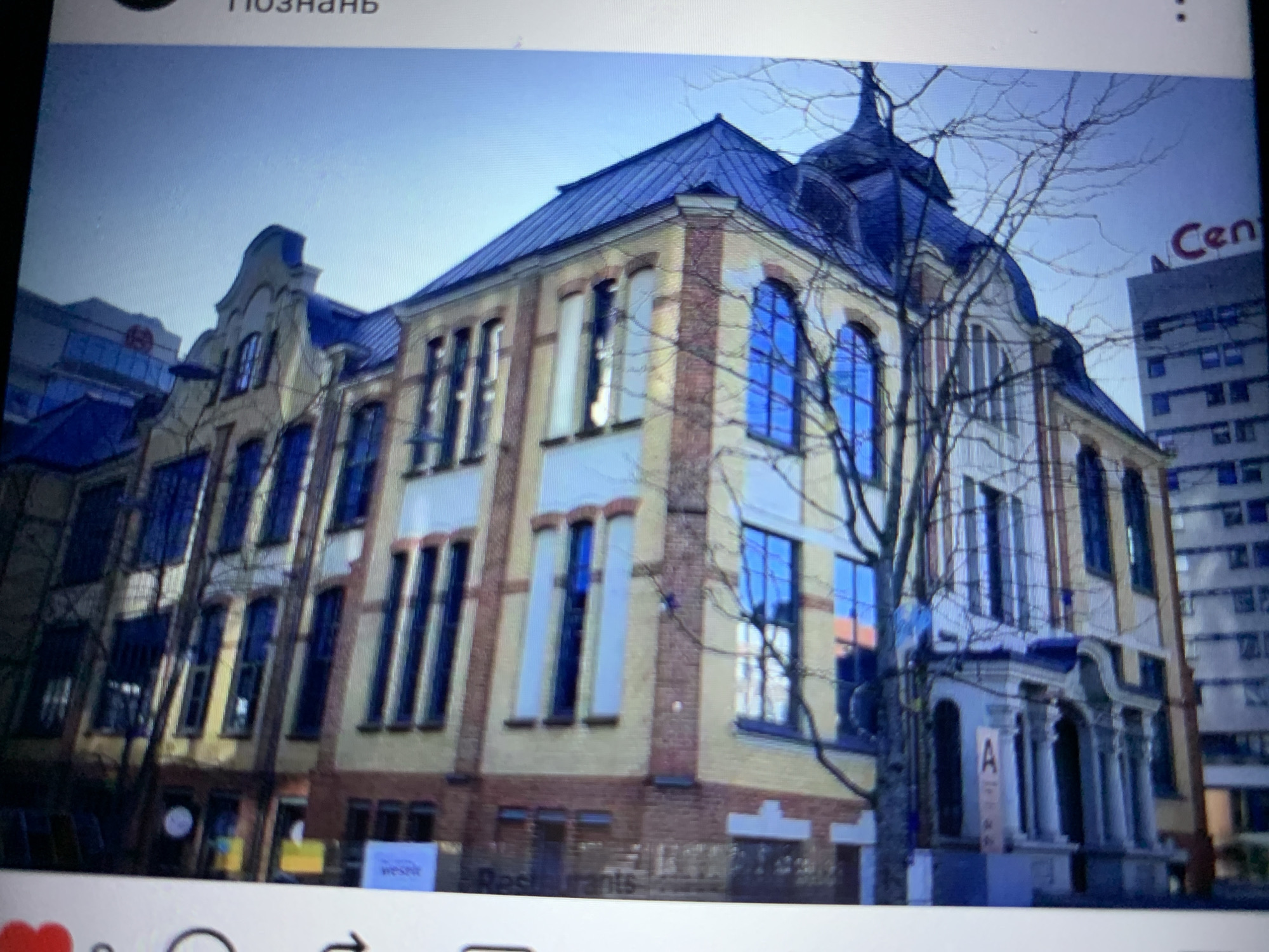 The image is a photograph of a building with a distinct architectural style, possibly a historical or renovated structure. The building features multiple stories with numerous windows, many of which are reflective and appear to be blue. The facade is a combination of light-colored material and red brickwork. A dark blue, angular roofline is visible on the upper portion of the building. Bare tree branches are in the foreground, obscuring parts of the building. The sky is a clear, pale blue. The photograph is taken at an angle, making the building appear tilted. On the right side of the image, a portion of text is visible vertically, reading "ПОЗНАНЬ" which translates to "KNOWLEDGE". There is also a partial view of another building in the background. The context of Lymanka, Ukraine is provided, but the image itself does not contain any direct visual cues to confirm this specific location.