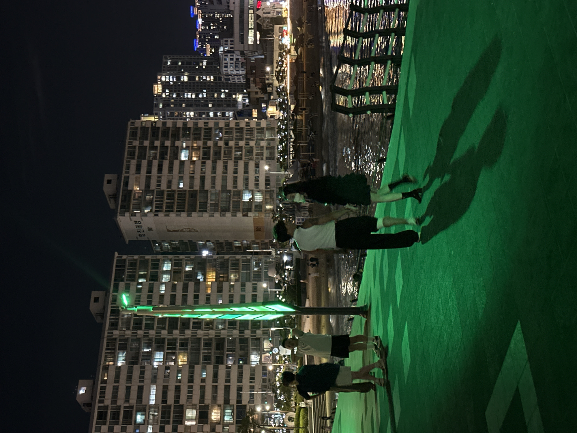 The image depicts a nighttime scene in Busan, South Korea, likely at the Haeundae Beach area. The foreground features a vibrant green walkway with geometric patterns. Several people are present on this walkway. In the center, a young couple stands facing each other, with the man wearing a white sleeveless shirt and black shorts, and the woman in a black dress and face mask. To their left, another person in a dark top and light-colored shorts stands facing away from the camera, and to the far left, another individual in a dark top and shorts is also visible. To the right of the couple, a person in a white tank top and black pants is seen from behind.
In the background, a tall lamppost with a striking green illumination stands prominently. Beyond the walkway, there is a body of water reflecting the lights from buildings and streetlights. A beach area with many sunshades and people is visible across the water, suggesting it is a popular gathering spot. Towering apartment buildings, illuminated with many windows, line the cityscape behind the beach, typical of urban coastal areas in South Korea. The overall atmosphere is one of urban nightlife at a seaside location.
