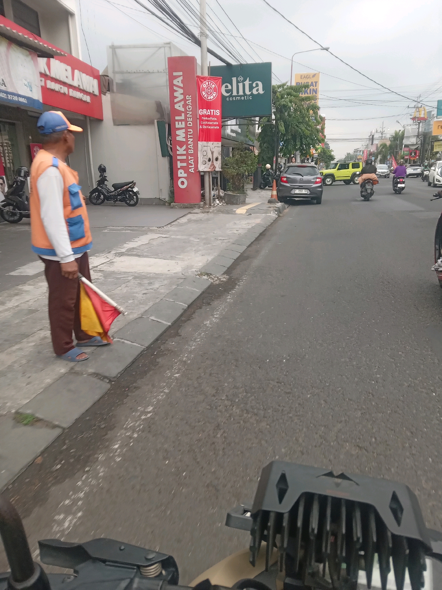 The image shows a street scene in Sinduadi, Indonesia. A man wearing an orange safety vest and a blue cap stands on a sidewalk, holding a red and yellow flag. He is facing away from the camera and to the left. In the background, there are several buildings with signs, including "OPTIC MELAWAI" and "ELITA COSMETIC." There are also motorcycles parked on the side of the road and vehicles driving on the street. The sky is overcast.