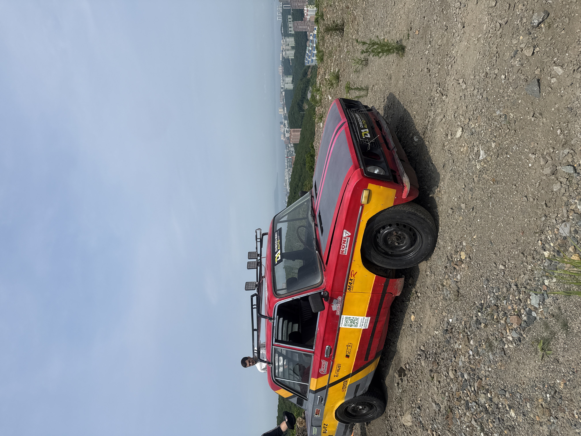 A modified red and yellow Lada sedan is parked on a gravelly slope overlooking a cityscape and ocean in Vladivostok, Russia. The car has a roof rack with auxiliary lights, various decals and sponsorships, and appears to be prepared for off-road use or racing. Two individuals are partially visible near the car. One person is standing by the open driver's side door, and another person's legs are visible on the left side of the frame, appearing to be sitting on a rock. The background shows a dense urban area with many buildings, including high-rises, nestled among green hills that slope down towards a body of water, likely the Ussuri Bay. The sky is overcast with a light blue hue. The overall scene suggests a promotional event, an off-road excursion, or a rally preparation.