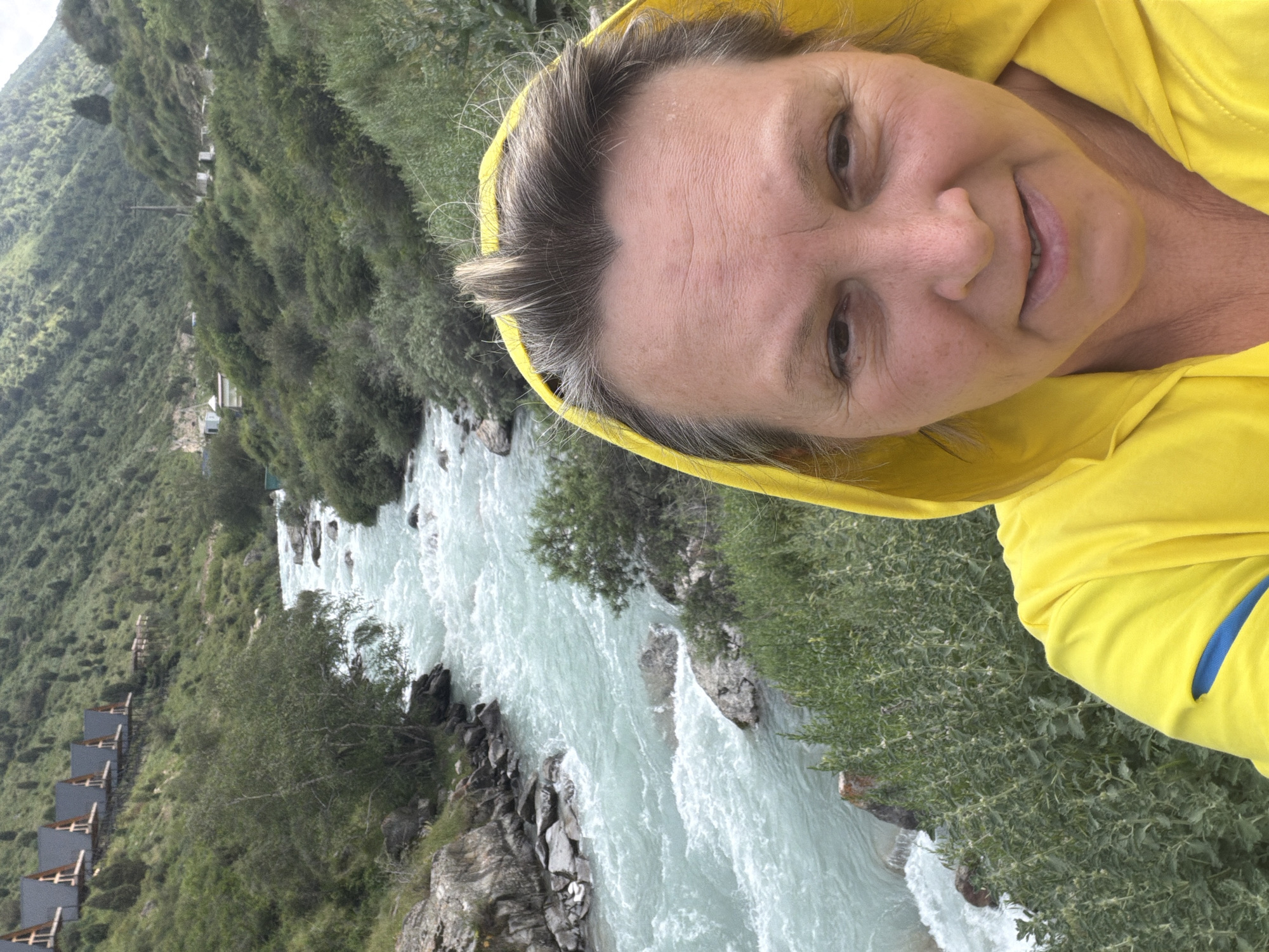 A selfie of a woman wearing a yellow hooded jacket is taken in front of a natural landscape. The woman's face fills the foreground, with her eyes looking towards the camera and a slight smile on her lips. Her hair is pulled back, revealing her forehead and temples. Behind her, a turquoise river flows rapidly through a rocky gorge. The banks of the river are lined with lush green vegetation and trees. In the distance, on the left side of the frame, a row of modern chalets with dark triangular roofs are visible, perched on a steep, tree-covered hillside. The overall scene suggests a remote, mountainous location.