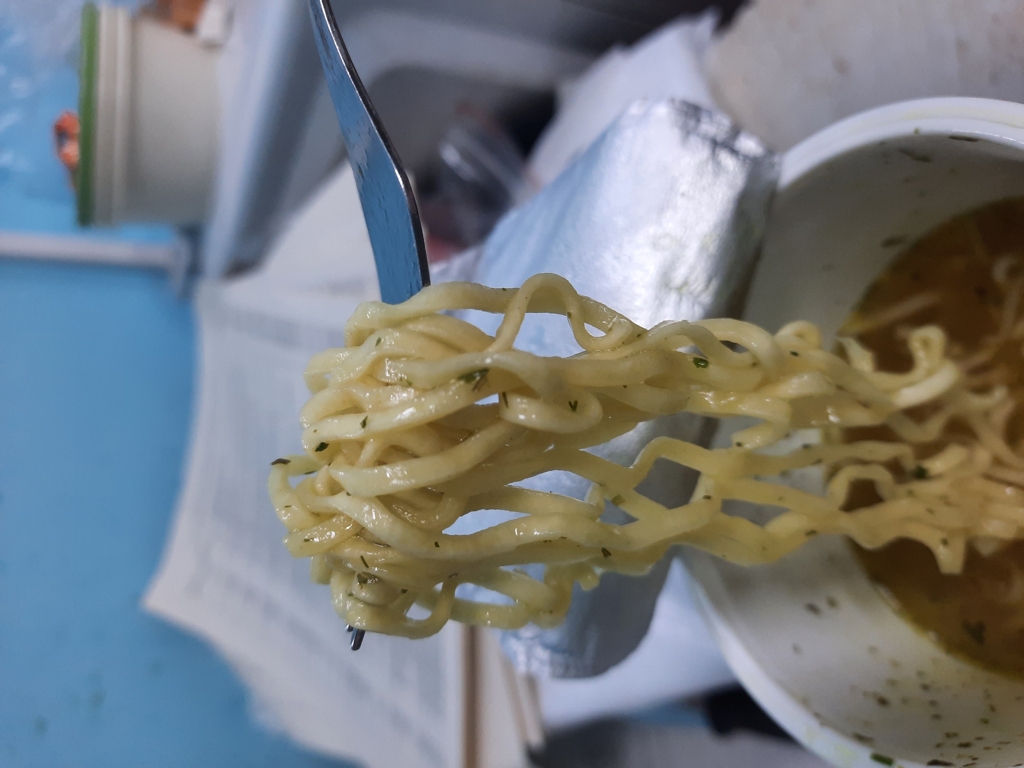 A fork holds a clump of noodles, with the container of noodles visible in the background. The noodles appear to be covered in a sauce or oil, possibly flavored with herbs. The background is cluttered with what appears to be packaging and a container with a green lid. The scene likely takes place indoors. There is no evidence to indicate that the picture was taken in the city of Krasnogorskoye, Russia. The only information that is available is regarding noodles on a fork in front of a container of noodles.