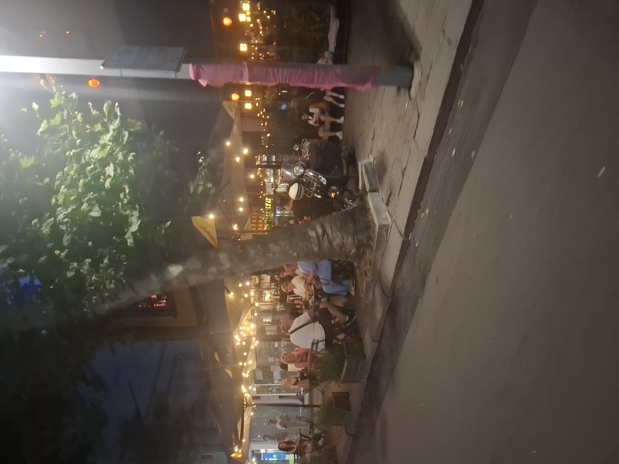 The image shows an outdoor seating area at night, likely a restaurant or cafe. Several people are seated at tables, engaged in conversation and enjoying their time. String lights illuminate the scene, creating a warm and inviting atmosphere. A motorcycle is parked nearby, adding to the urban ambiance. The time of day is clearly night, with artificial lights providing the primary illumination. The weather appears to be pleasant, as people are seated outdoors. There are no specific location cues visible in the image. The overall mood of the scene is relaxed and social. No visible text is present.