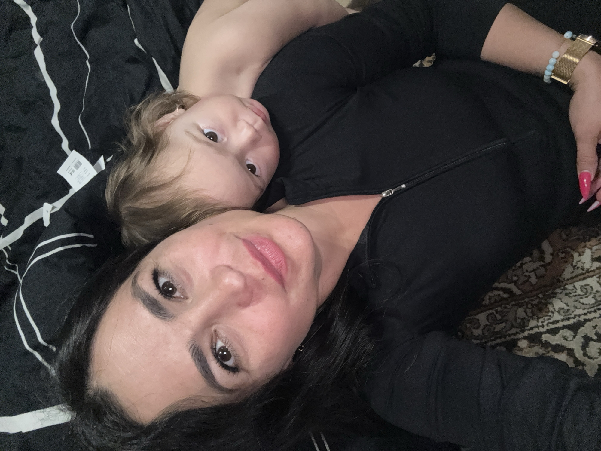 This eye-level indoor photo captures an intimate moment between an adult woman and a young child, presumably a mother and child, in Chaikovsky, Russia. Both individuals are lying down, looking directly towards the camera.
The woman is in the foreground, characterized by her long dark hair, dark eyes, and a soft, gentle smile. She is wearing a black long-sleeved zipped top. Her left wrist displays a gold-toned watch and a light blue beaded bracelet, and her long fingernails are painted a vibrant pink.
The child, with light brown hair and dark, curious eyes, is positioned slightly behind the woman, resting their head on her shoulder or chest. The child's expression is neutral yet engaged, looking towards the camera.
The setting appears to be a cozy indoor environment. On the left, a black fabric surface with prominent white stripes, likely bedding or a blanket, is visible. A small white product tag with black, illegible text is attached to this fabric near the child's head. On the right side of the frame, a segment of a patterned rug or carpet, featuring intricate designs in shades of brown, beige, and black, adds to the homey ambiance. The lighting is soft and even, suggesting a relaxed atmosphere. The overall mood of the image is one of warmth and affection.