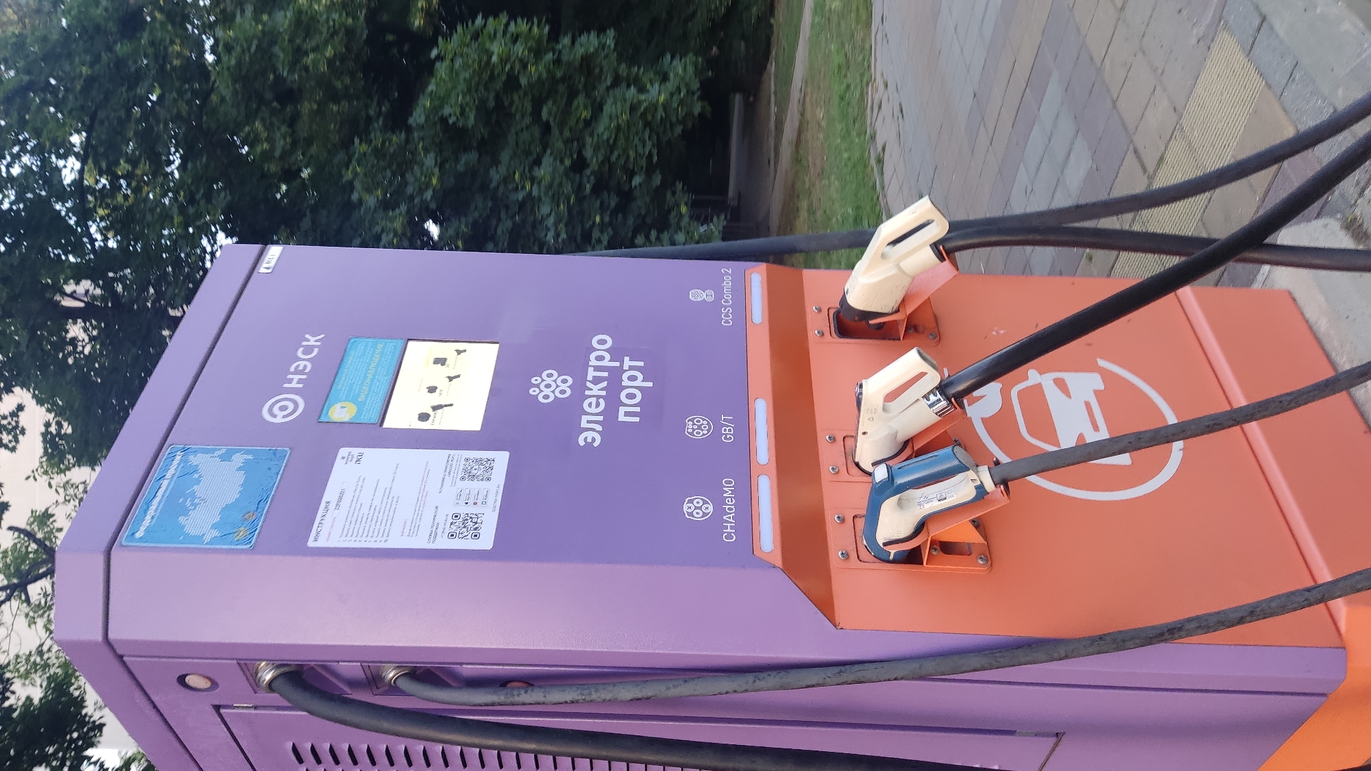 An outdoor electric vehicle charging station, identified by the logo "Электро порт" and "НЭСК," stands on a paved area with trees in the background. The charging station is predominantly purple and orange. It features three charging connectors: one blue and white, and two beige, all connected to cables. Above the connectors, the station displays information, including a map of Russia, instructions, and logos for CHAdeMO, GB/T, and CCS Combo 2 charging standards. A white silhouette of a car is visible on the orange section of the station. The scene suggests a public space in Krasnodar, Russia, likely a street or parking area, given the presence of trees, pavement, and the charging infrastructure. No people are present in the image.