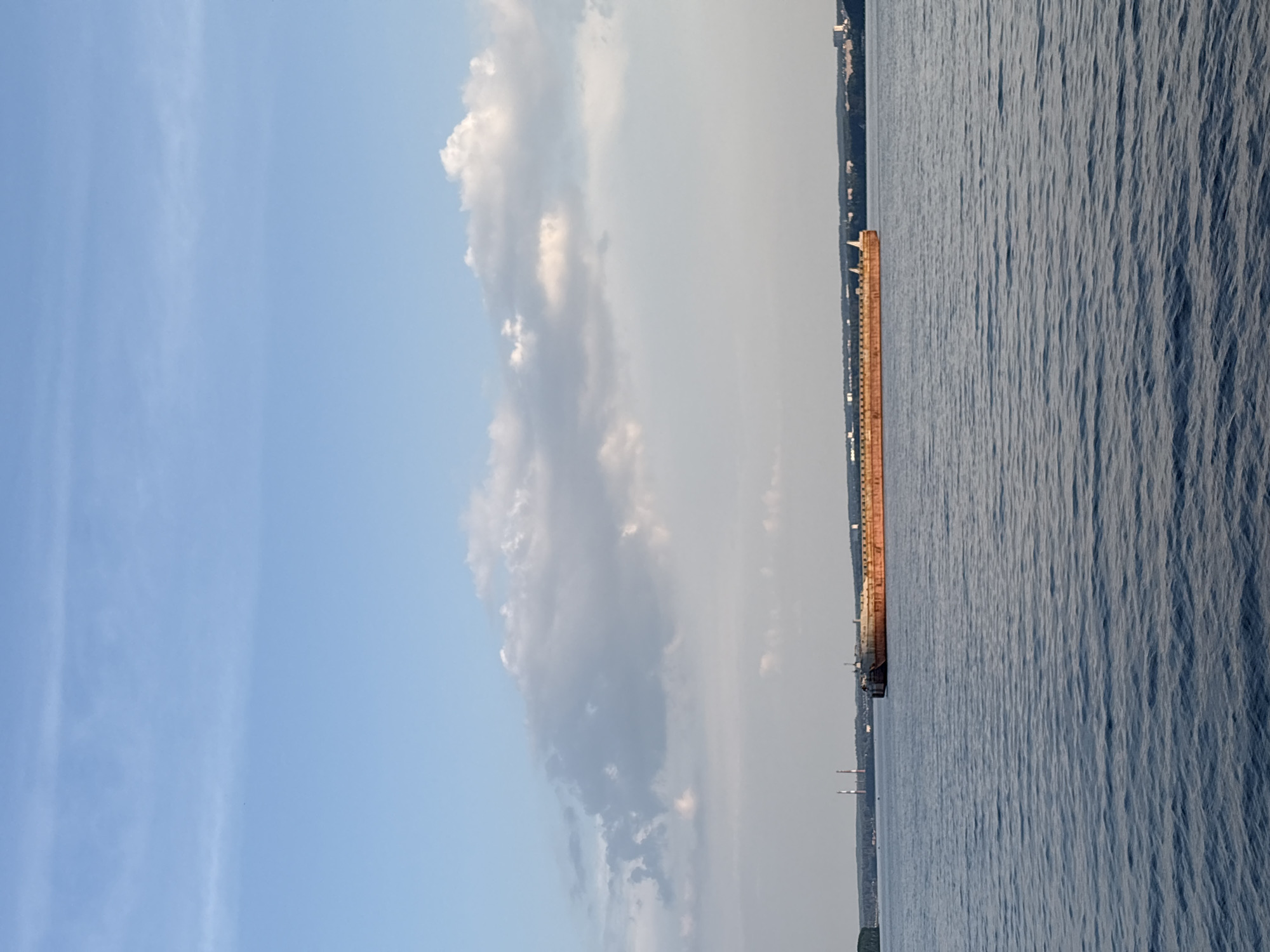 The image shows a wide, calm body of water, likely a river or reservoir, under a pale blue sky with scattered white clouds. In the distance, a long, flat-topped barge, painted a reddish-brown, is moving across the water from left to right. The barge is heavily laden, its deck lined with vertical structures.
On the far bank of the water, a treeline is visible, with several buildings and two tall, slender industrial smokestacks with red and white bands. The landscape suggests an industrial area. The water's surface is rippled, indicating a gentle breeze. The overall scene is serene, with the barge being the dominant man-made object in the natural environment. This scene is consistent with the appearance of the Kama River near Chaikovsky, Russia, a city known for its industrial sector and hydroelectric power station, which would explain the presence of the barge and smokestacks.