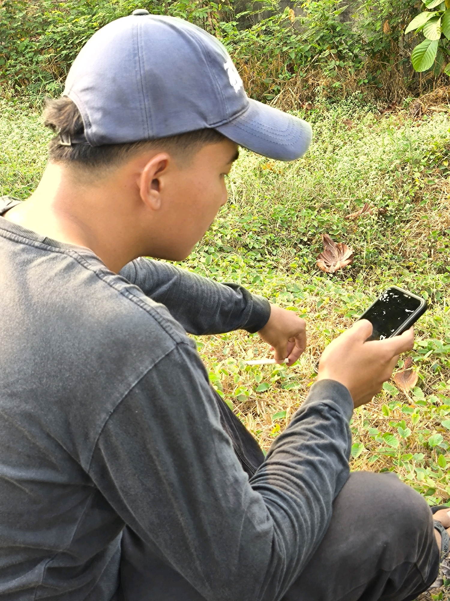 In a grassy outdoor area in Mekarwangi, Indonesia, a person wearing a gray long-sleeved shirt and a blue baseball cap is seated. The person is holding a smartphone in their right hand and appears to be interacting with it. A white cylindrical object, possibly a cigarette or pen, is held in their left hand. The background consists of lush green vegetation and dry leaves scattered on the ground. The lighting suggests it is daytime.