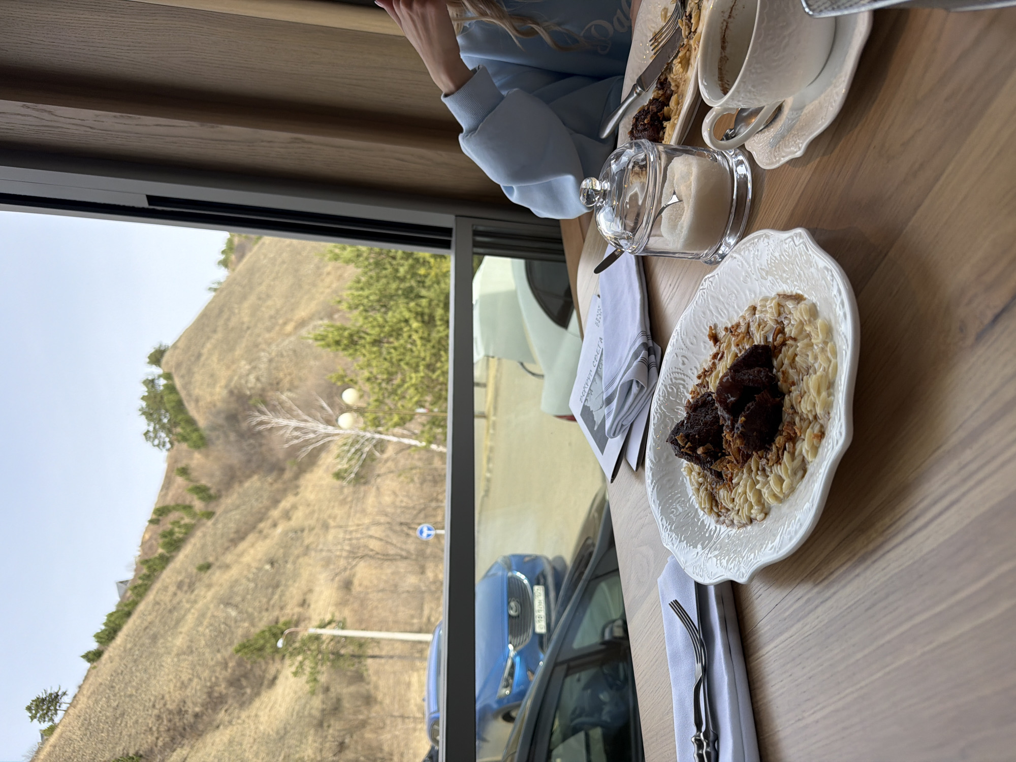The image captures a dining scene near a window in what appears to be a restaurant or cafe. The view from the window shows a hilly landscape with sparse vegetation, suggesting a location in or near a natural setting. A car is parked outside.
On the table, a person wearing a light blue top is seated. There are plates of food, a sugar bowl, and a cup and saucer. Silverware is neatly arranged on a white napkin. The focus is on the food, which appears to be a savory dish.
Given the mention of the city in the request, it's probable that the image was taken somewhere in or near Krasnoyarsk, Russia, where the landscape and automotive traffic align. The image shows a blend of indoor dining and natural surroundings.