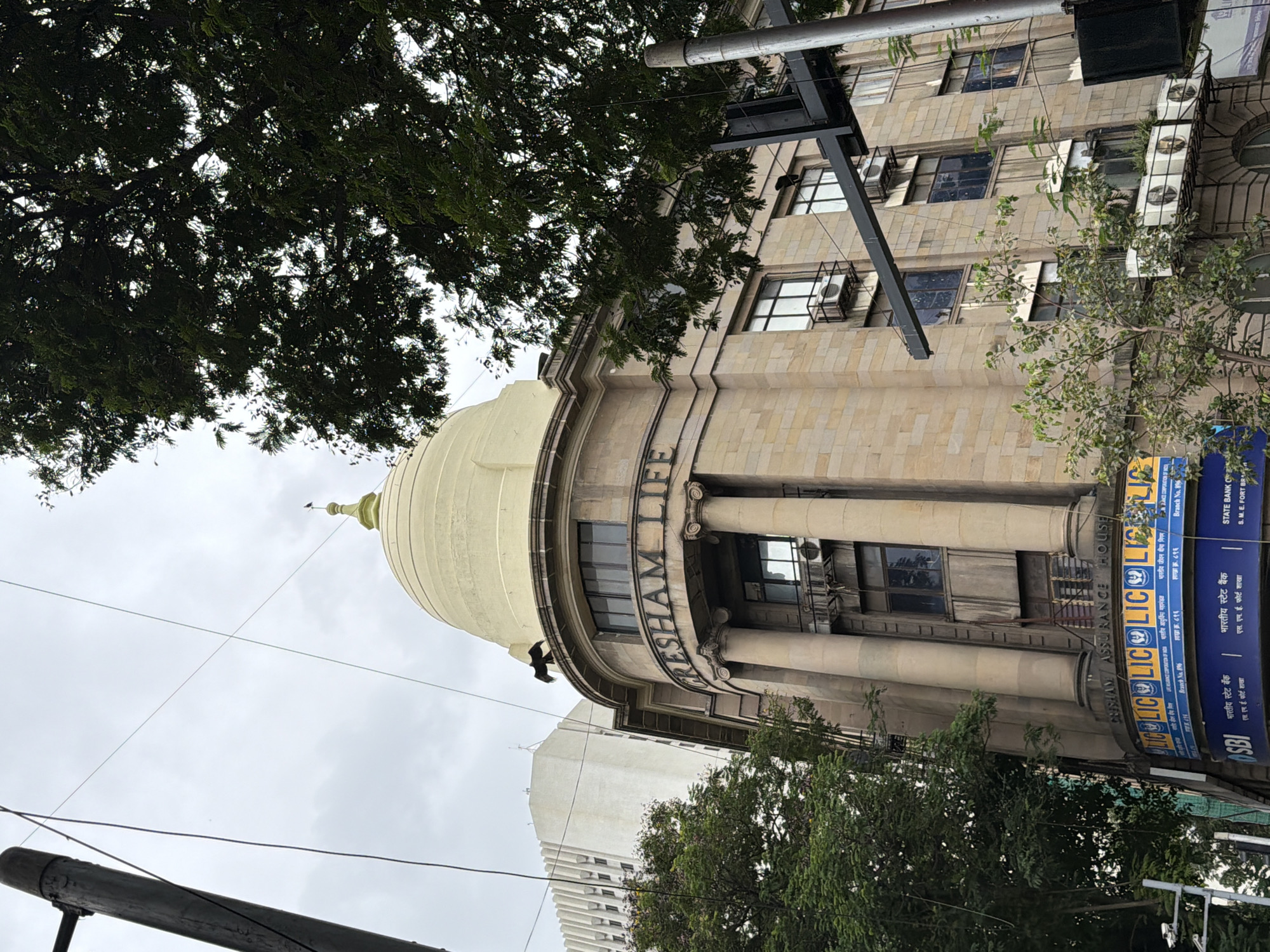 A low-angle, exterior, daytime, medium shot captures the Gresham Assurance House, a historical building in Mumbai, India, under a cloudy sky. The building, constructed with light-brown stone, features a distinctive golden dome. The name "Gresham Life" is prominently displayed in large, dark letters above a portico supported by tall, fluted columns. Below the portico, a curved facade is adorned with the blue and yellow "LIC" logo, indicating the Life Insurance Corporation of India. The SBI (State Bank of India) logo is also visible on the lower portion of the building.
In the foreground, to the left, a dark wooden utility pole stands tall, draped with multiple power lines. A large, dark green tree with dense foliage partially obscures the upper right portion of the Gresham Assurance House and extends towards the top right corner of the frame. On the dome of the building, a dark bird is perched, facing left. To the left of the Gresham Assurance House, a taller, modern white building with a grid-like facade is visible. The image also shows several windows with air conditioning units mounted on the exterior. A street light pole and a security camera are visible on the lower left side. The overall scene depicts an urban environment with a blend of historic and modern architecture, set against an overcast sky.