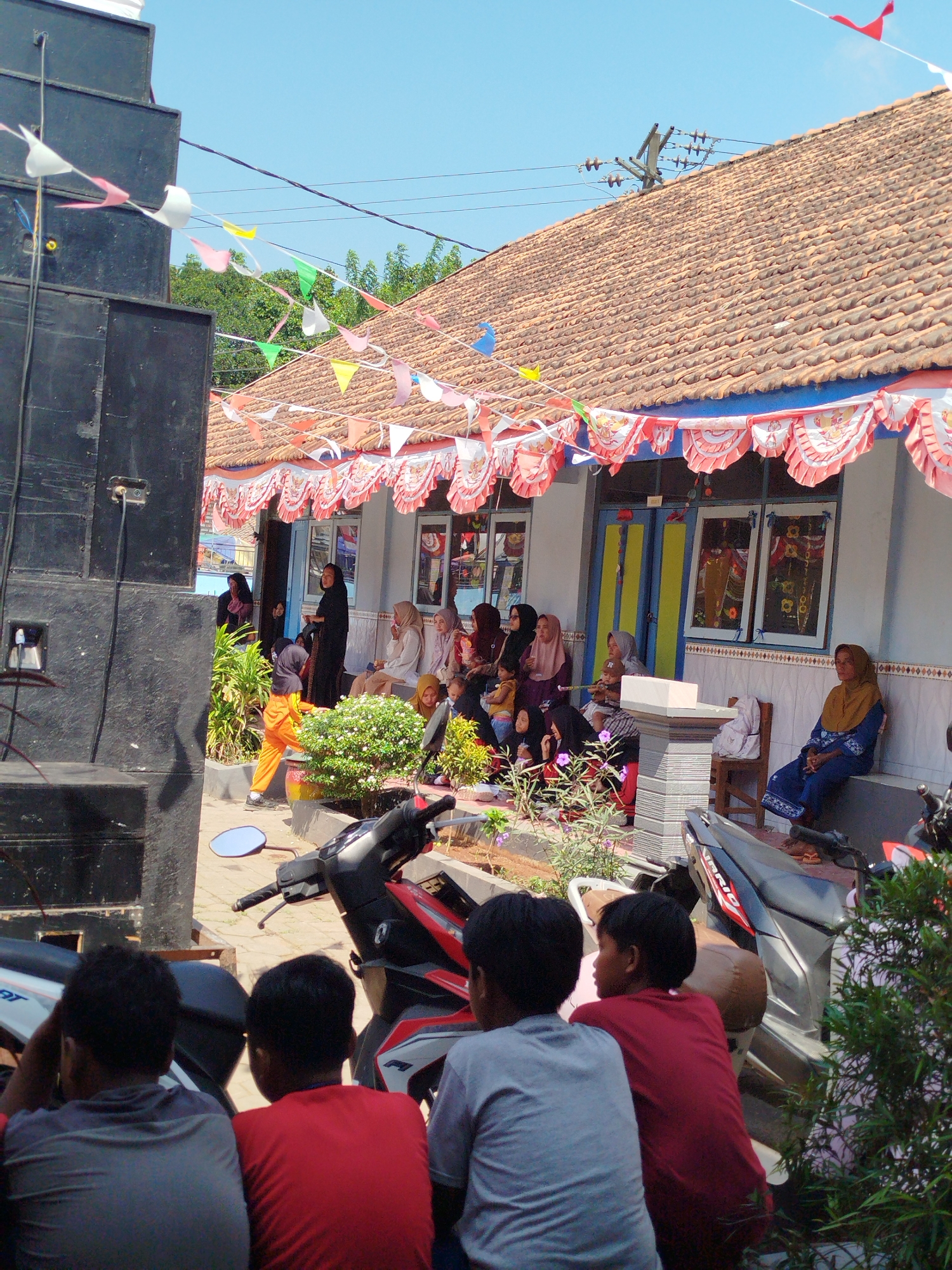 A group of people are gathered outside a building in Parseh, Indonesia, decorated with red and white bunting. On the left, a large black speaker tower is positioned. Several individuals, including women and children, are seated and standing near the entrance of the building, which has tiled roofing and windows. A young person in orange pants is walking towards the building. In the foreground, the backs of three young males are visible, seated behind a motorcycle. Red and green triangular flags are strung across the scene. The environment appears to be an outdoor gathering or event.