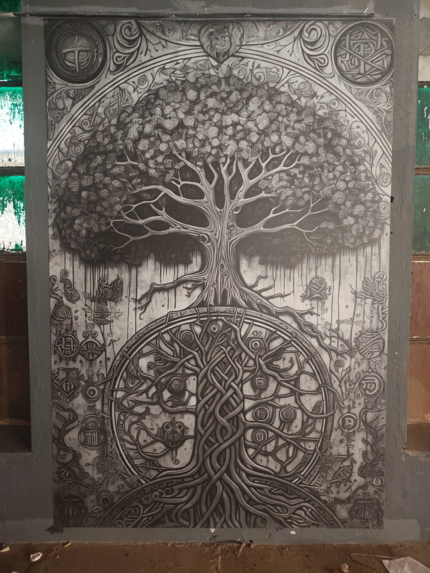 The image shows a large, detailed charcoal drawing of a tree, possibly the Tree of Life, affixed to a wall. The tree has a broad canopy of textured leaves and exposed roots which form a circular pattern below. The artwork is surrounded by a border featuring Celtic knotwork designs and stylized faces. The wall appears to be part of an old or unfinished building, with a window visible on the left side. The wall is painted in a grey-blue color and is positioned at the city of Kalamassery, India. The ground is brown and littered with debris.