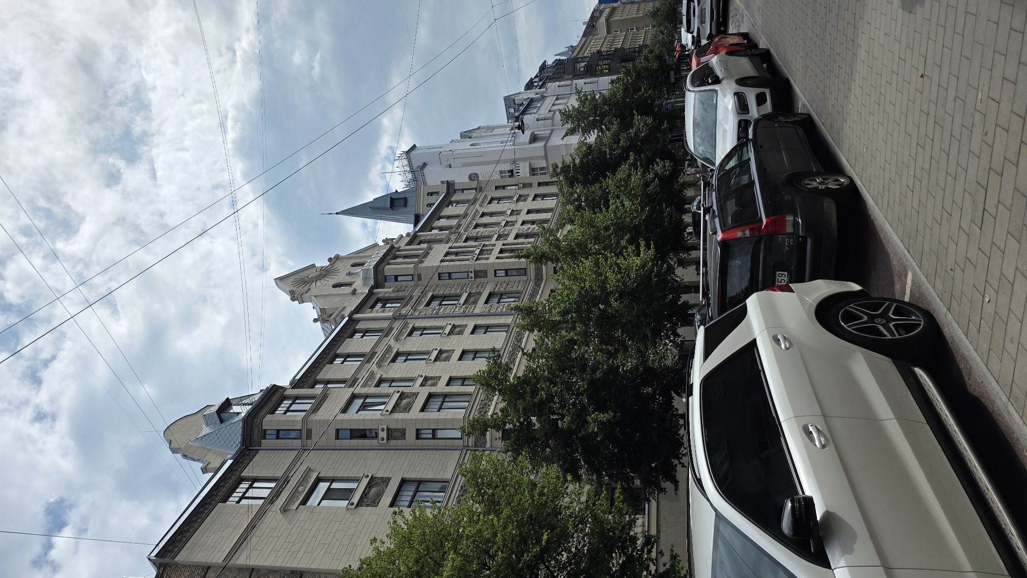 The image captures a street scene in Riga, Latvia, dominated by a row of ornate Art Nouveau buildings lining a cobblestone sidewalk. The architecture features intricate facades, mansard roofs, and decorative elements. Lush green trees are planted along the sidewalk, separating the buildings from the street. A line of parked cars is visible on the street, including a white Mercedes-Benz SUV in the foreground, a black Volvo wagon, and other vehicles further down the road. Overhead, telephone or power lines crisscross the cloudy sky. The overall impression is one of a well-preserved urban environment, showcasing Riga's distinctive architectural heritage.
