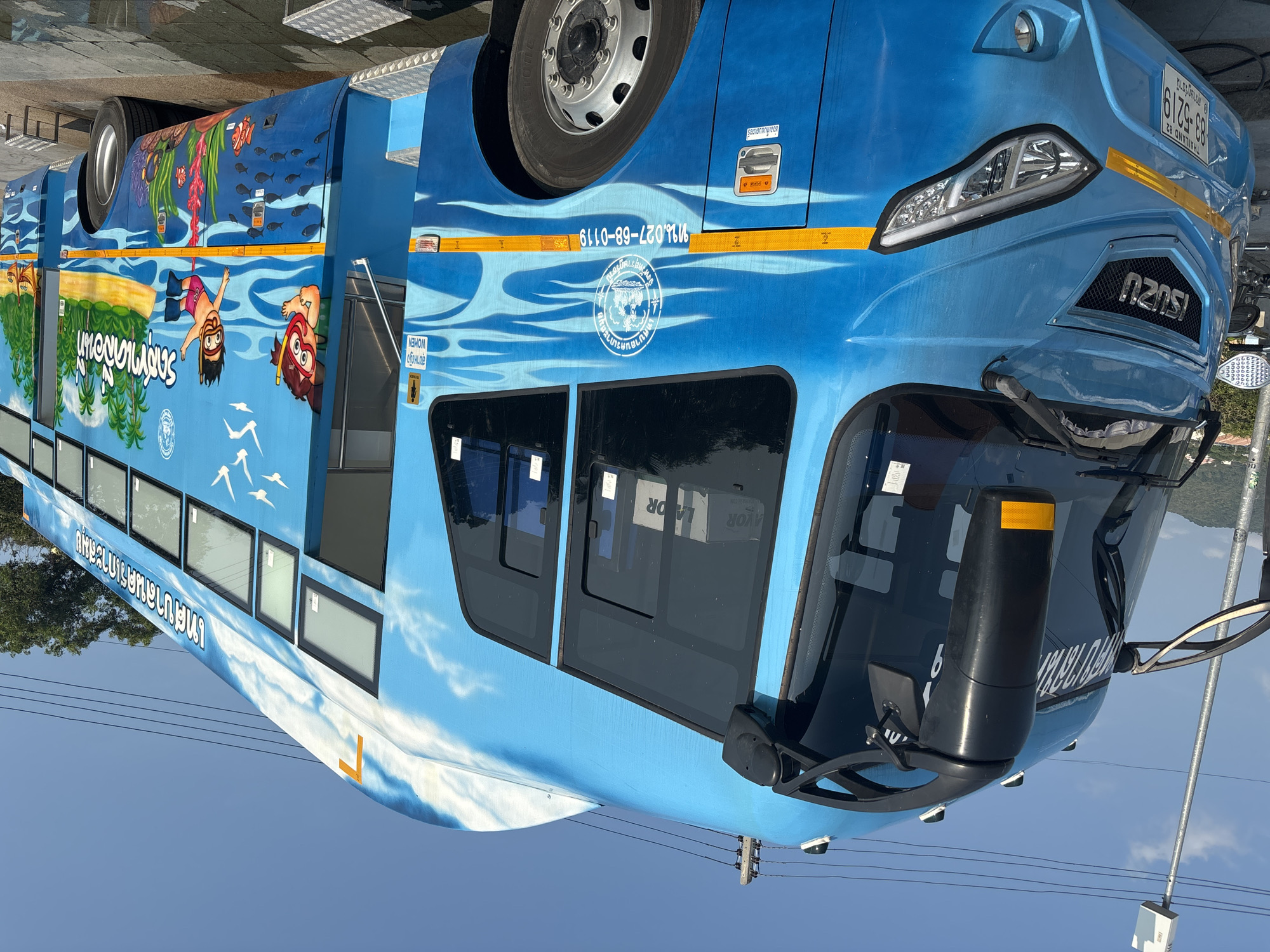 The image shows a blue ISUZU bus, likely located in บ้านบางรักษ์, Thailand, based on the license plate information, specifically "THAILAND 82" and "สุราษฎร์ธานี" (Surat Thani), which is the province where บ้านบางรักษ์ is located. The bus is decorated with ocean-themed artwork, including depictions of divers, tropical fish, and underwater scenery. Markings indicate it belongs to the "เทศบาลนครเกาะสมุย" (Koh Samui Municipality). A phone number "ทน.027-68-0119" is displayed. The bus features labels such as "WOMEN" for the restroom (สุขาหญิง) and "ช่องเก็บแบตเตอรี" (battery storage). The license plate reads "83-5219." The bus is parked on a paved surface, with utility poles and power lines visible in the background. The sky is a light blue color.