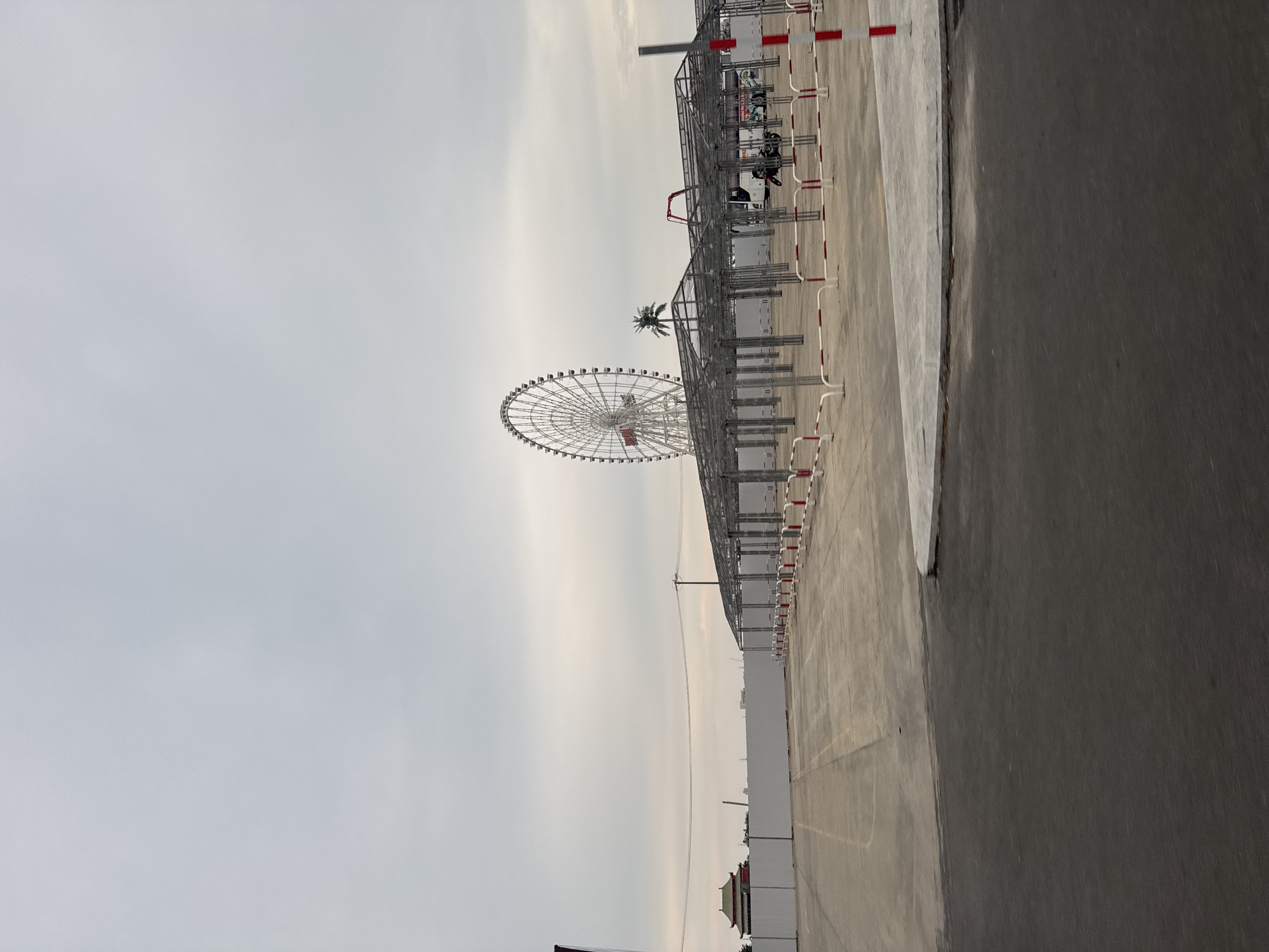 The image depicts a Ferris wheel in Đà Nẵng, Vietnam, seen from an elevated perspective within what appears to be a parking or staging area. A partially constructed metal framework with red and white safety barriers extends along the right side of the frame. In the background, a building or structure with a traditional Vietnamese-style roof can be seen. The sky is overcast.