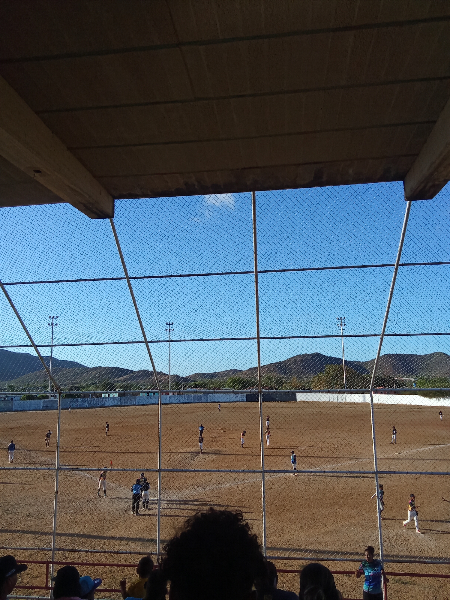 The image shows a baseball field in Juan Griego, Venezuela. The view is from a covered seating area behind a chain-link fence. Players are dispersed across the dirt field. In the background, there is a wall or fence, light posts, and rolling hills under a blue sky with a single cloud. People can be seen in the foreground, either in the seating area or walking near the field.