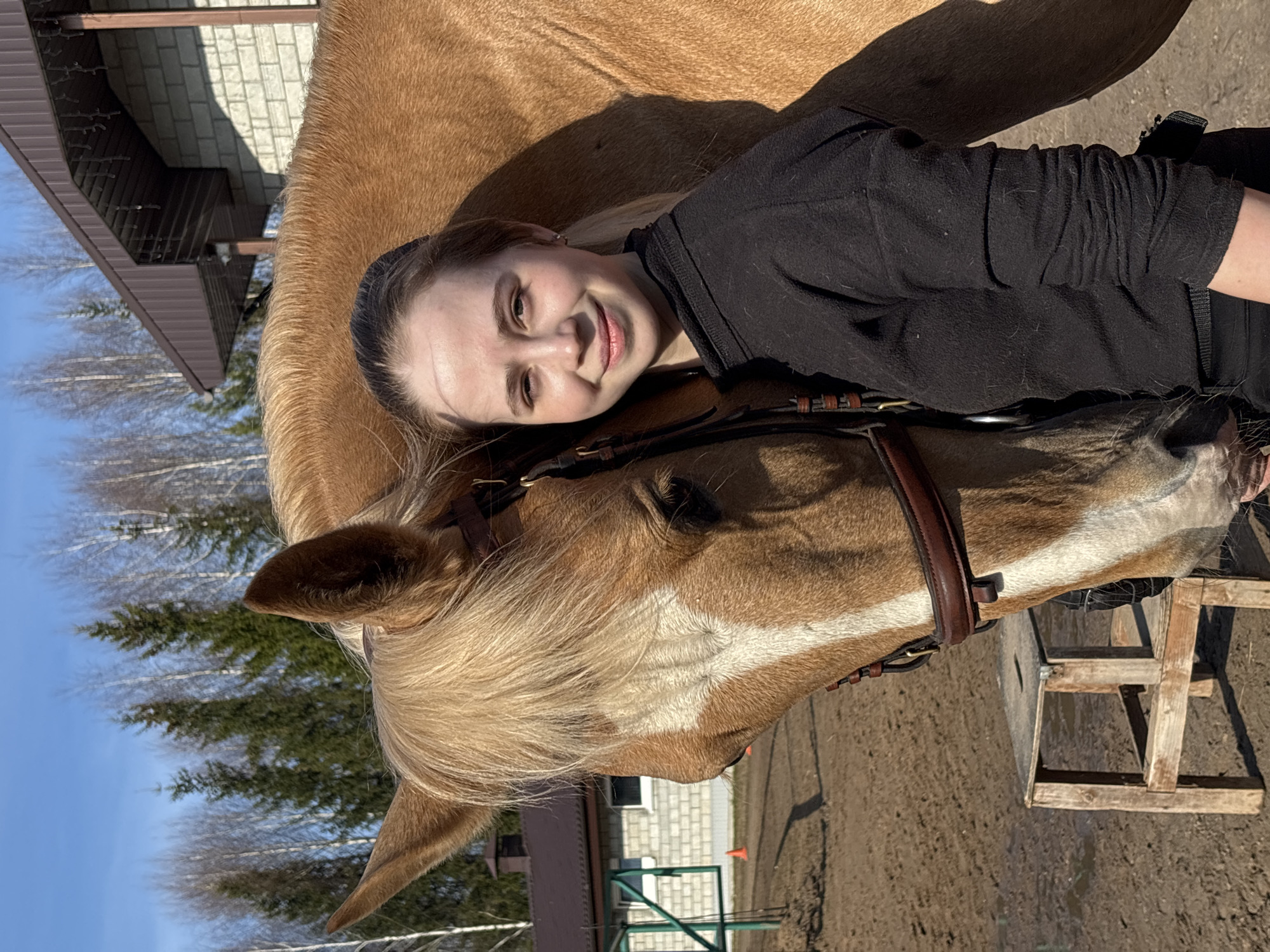 The image shows a fair-skinned young woman with blonde hair and a black long-sleeved shirt, lying her head on the head of a light brown horse. The horse is wearing a bridle. In the background are trees, buildings, and what appears to be an outdoor training area for horses. While the prompt specified the city of Русский Вожой, Russia, it's impossible to verify this information from the image alone. Therefore, I cannot definitively state the location is Русский Вожой, Russia based solely on the visual content of the provided image.