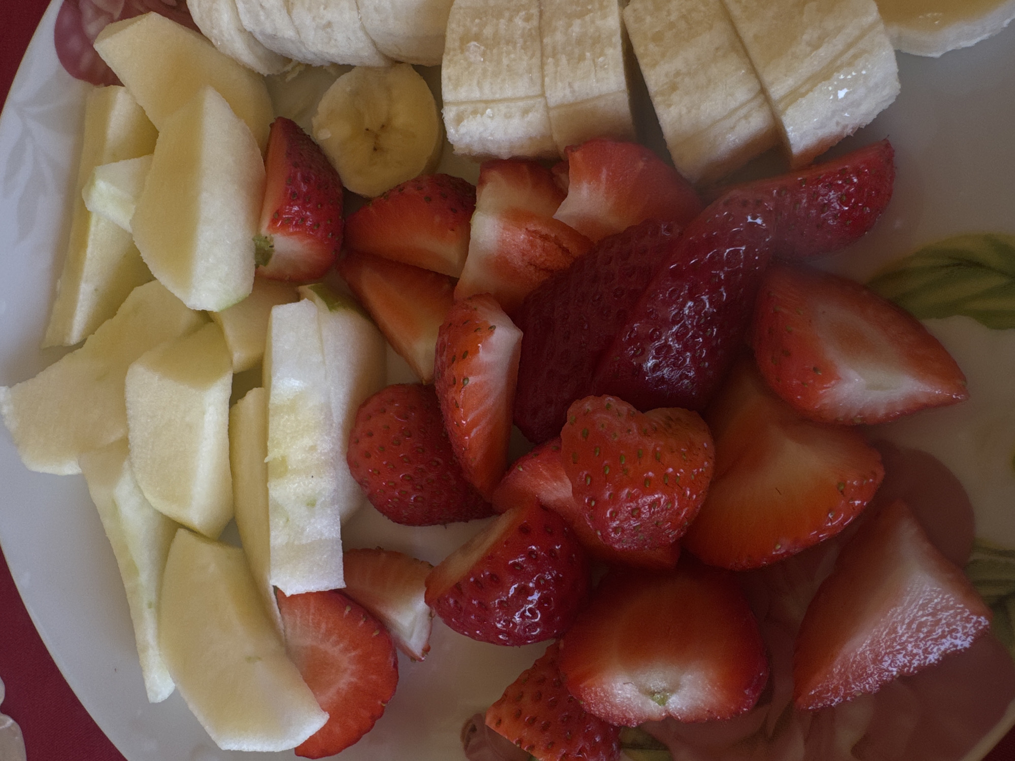A close-up overhead shot shows a plate of sliced fruit, including apples, bananas, and strawberries. The apples are cut into rectangular segments, the bananas into round slices, and the strawberries mostly halved or quartered. There are no people visible. The plate appears to be ceramic and rests on a red surface. The location is not indicated and not discernable from the image.