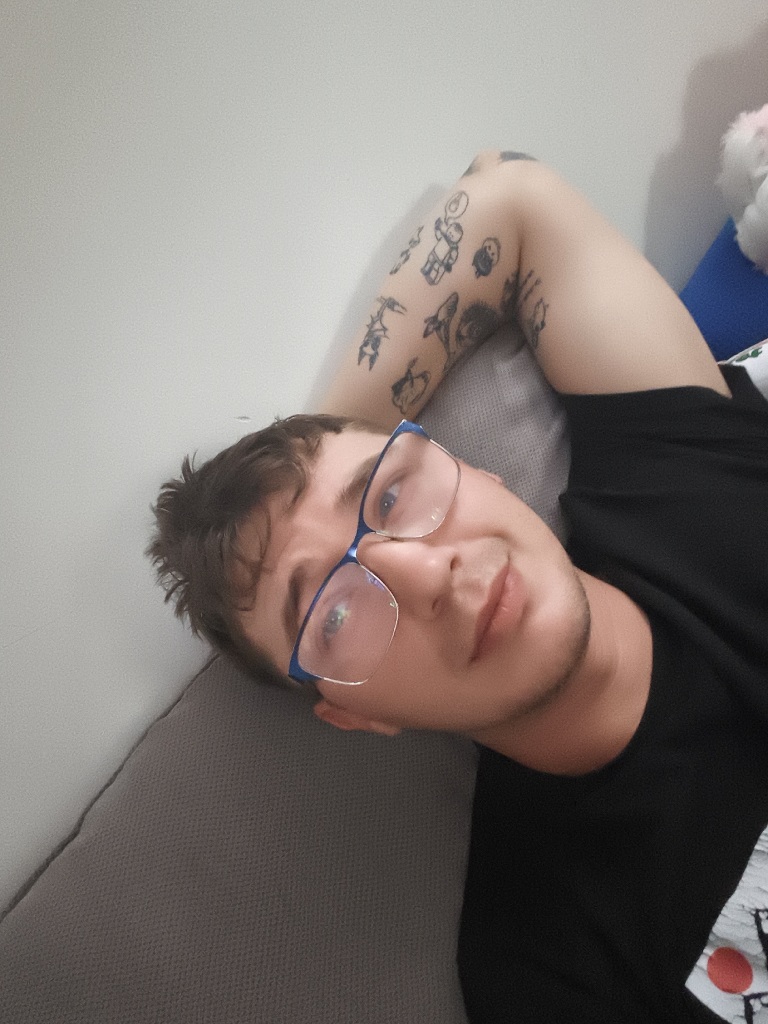 A fair-skinned man with short brown hair and a light beard is lying on a gray couch with his head tilted towards the camera. He is wearing blue-framed glasses. His left arm is raised and bent, resting behind his head. The arm features several black ink tattoos. He is wearing a black t-shirt. Behind him is a white wall. To the right, part of a blue pillow and a white object are visible. This scene is likely inside a residential dwelling. Location in Nowy Tomyśl, Poland cannot be determined from the image.