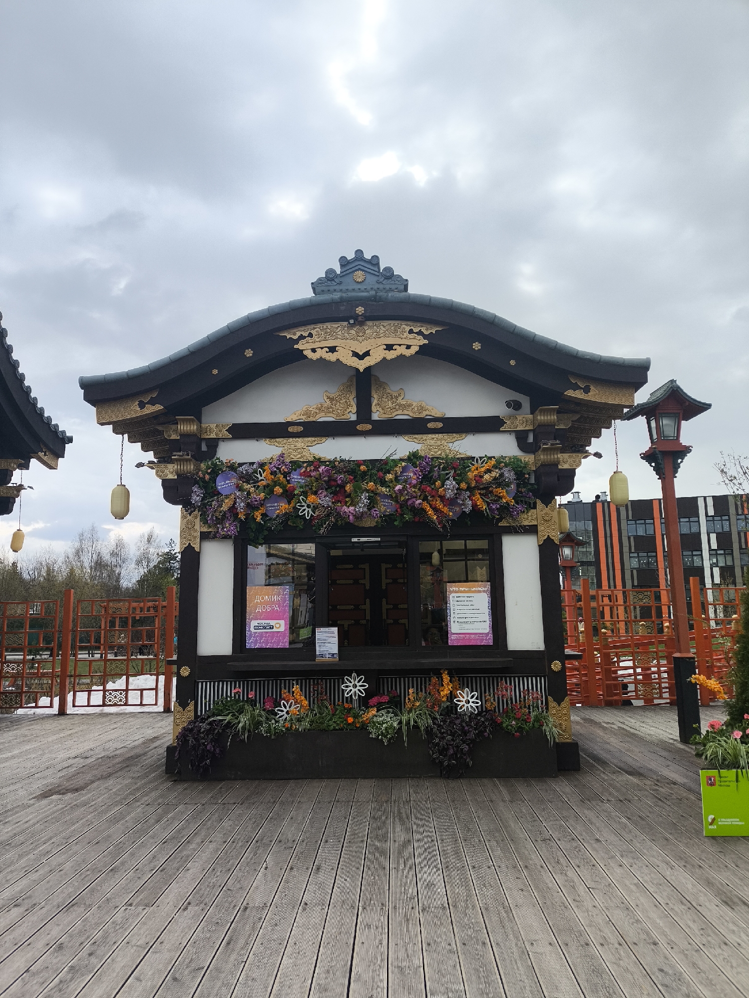 The image displays a small, elaborately decorated building in an outdoor setting, designed in a Japanese architectural style, situated on a wooden deck in Moscow, Russia. The building features white walls and a dark, curved roof embellished with intricate gold detailing and decorative elements. A prominent, vibrant arrangement of artificial flowers in purple, pink, orange, and yellow hues adorns the facade above the main window.
Several yellow, cylindrical lanterns hang from the eaves of the main building and an adjacent structure on the left. A traditional red Japanese-style lantern on a tall post is visible to the right. Below the main window, planter boxes are filled with various flowers and foliage, some artificial, alongside white decorative flower shapes. The ground is covered by a wide wooden deck made of horizontal planks. Red or orange lattice-style fencing, reminiscent of Japanese garden designs, is visible on both sides of the building.
In the background on the left, bare trees indicate a cooler season. To the right, modern, multi-story buildings with distinctive orange and dark striped facades are visible.
A sign on the left window of the building prominently displays "ДОМИК ДОБРА" (House of Good) followed by "ЧАСЫ РАБОТЫ:" (Opening Hours:), suggesting it functions as a kiosk or community service point. Another sign on the right window contains additional information. On a green planter or bin on the far right, partially visible text includes "Департамент культуры Москвы" (Department of Culture of Moscow) and phrases like "с соблюдением социальной дистанции" (observing social distancing), indicating a public space managed by the city.
The scene is captured during the daytime under an overcast sky, with no direct sunlight. No people are visible, contributing to a tranquil atmosphere.
