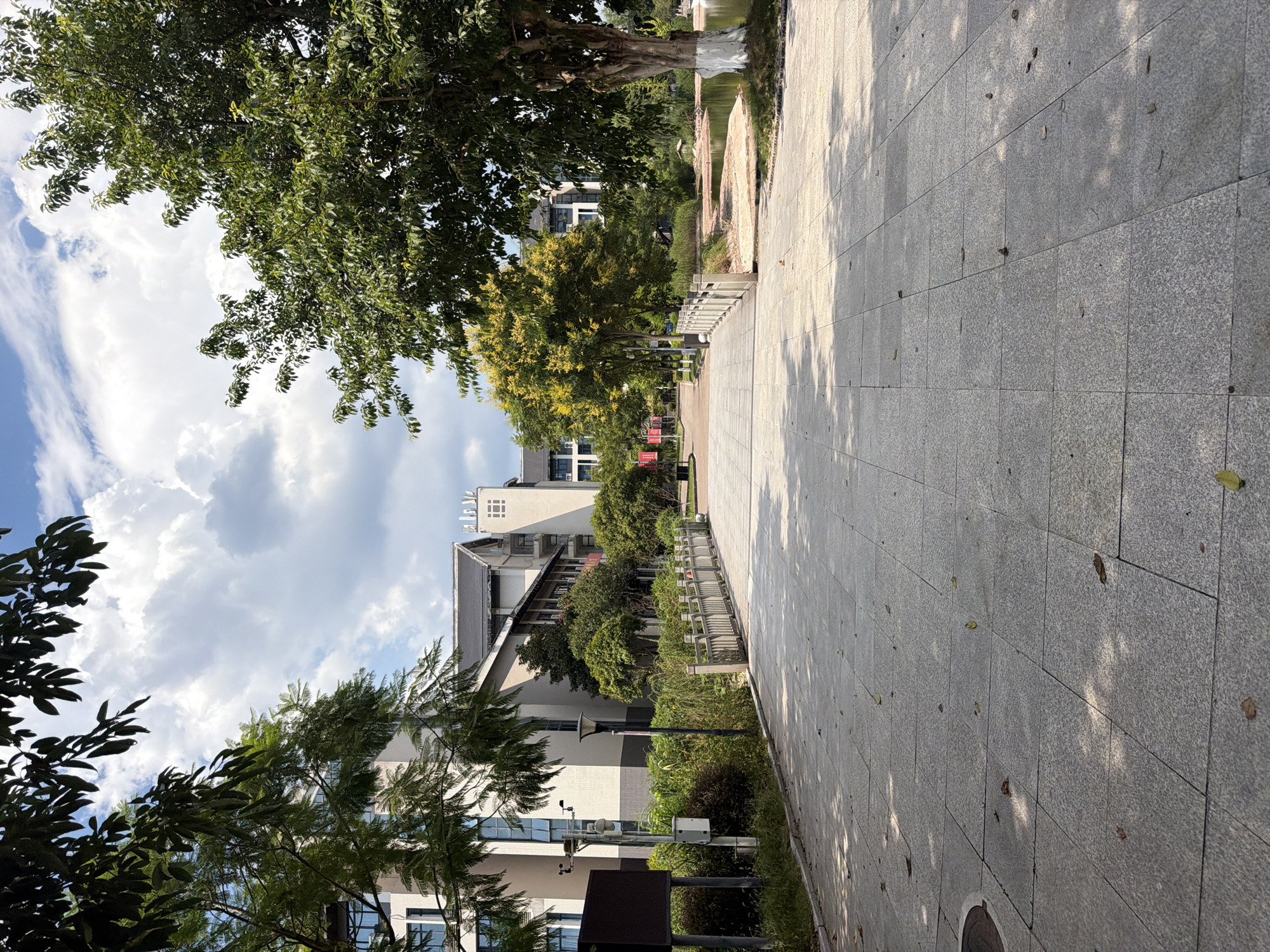 The image depicts a paved walkway leading towards a footbridge with a wooden railing. On the left side of the walkway, there are buildings with a modern architectural style, featuring multiple windows and light-colored facades. Lush green trees and vegetation line the walkway and surround the buildings. On the right side of the walkway, a body of water is visible, bordered by sandy banks and more trees. The sky above is partly cloudy with scattered white clouds against a blue backdrop. The walkway is illuminated by sunlight, casting shadows from the surrounding trees. The overall scene suggests an outdoor public space, possibly a park or a campus environment, in Jiangjin, China. There are no people or animals visible in the image.