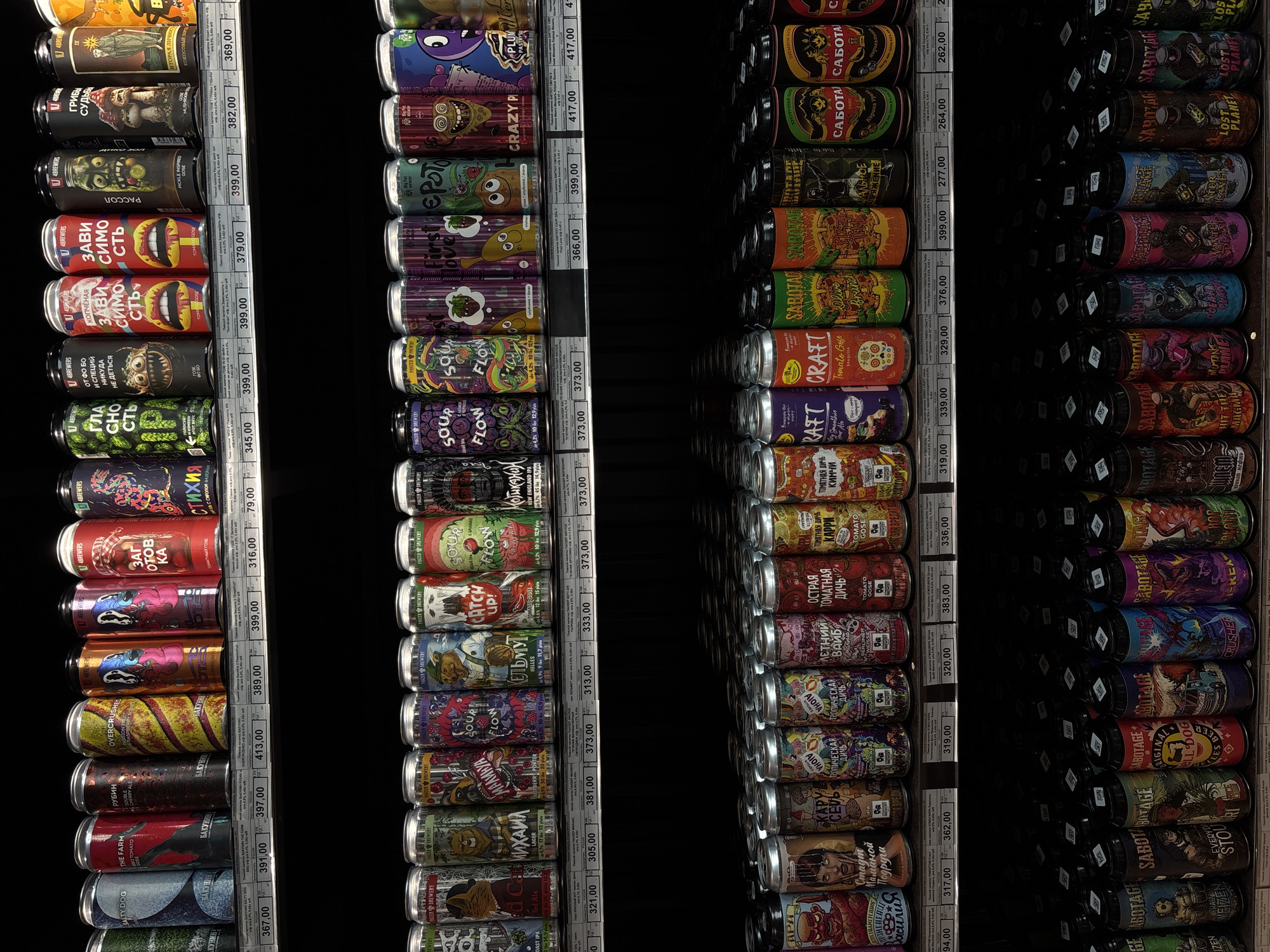 The image shows shelves stocked with canned beers. The shelves are vertically aligned, creating rows and columns of beers. Each beer can has a unique label, including cartoon illustrations, brand names (e.g., "4 BREWERS," "PANZER BREWERY"), and price tags.
The environment is darkly lit, focusing attention on the beer cans. The scene appears to be inside a shop or market, likely a section dedicated to craft beers. Price tags, such as "367,00", are visible. The labels include information about the beers, such as flavors, ingredients, and brewing details (e.g., "Tomato Gose," "Red Ale," "OAST IPA").