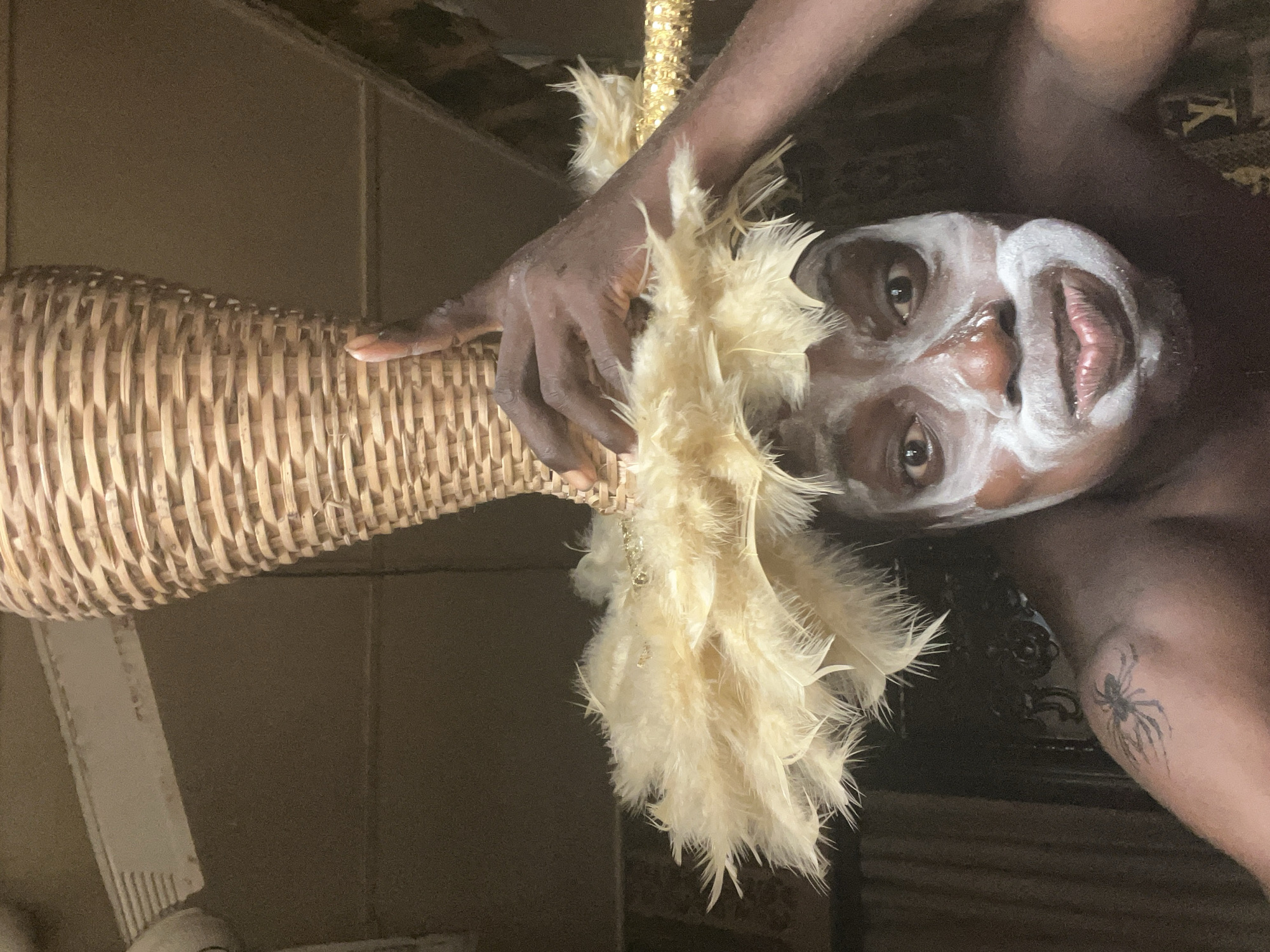 An adult individual with dark skin is captured in a close-up, sideways-oriented portrait, set indoors against a dark, indistinct background in Nkpor, Nigeria. The person's face is adorned with striking white markings, possibly paint or makeup, outlining the forehead, areas around the eyes and cheekbones, and encircling the mouth, leaving the lips and central facial features exposed. Their dark eyes look directly at the viewer with a neutral expression.
The person's right hand grasps the neck of a tall, light brown, cylindrical wicker vase. A cluster of fluffy, cream-colored feathers, possibly part of a decorative item or costume, drapes over their hand and the top of the vase. A shimmering golden element, possibly beads or sequins, is visible among the feathers near the hand. On the person's left upper arm, a distinct black spider tattoo is visible. The background includes a dark wall and a patterned fabric or rug in the upper right corner. The lighting appears to be artificial, typical of an indoor setting. No discernible text is visible in the image.