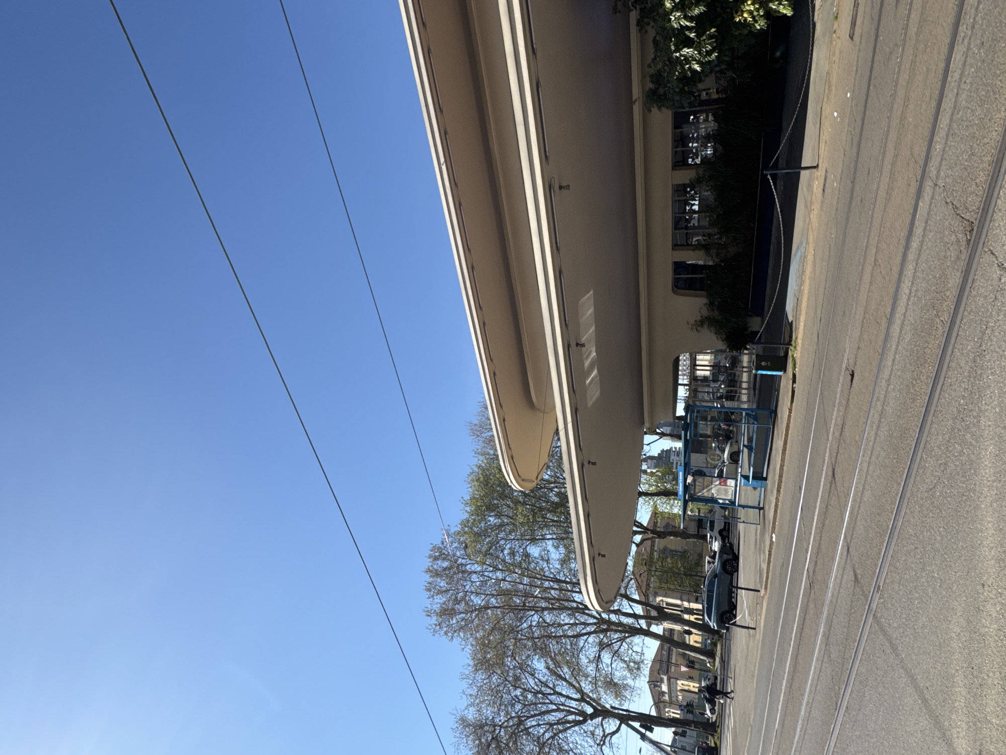 The image shows a street in Milan, Italy, featuring a tram line, a wide pavement, and a modern architectural structure overhead. The sky is clear and blue. There are overhead electrical lines. Trees line the street. Parked cars are visible along the sidewalk. The architectural structure is a curved, multi-layered building that casts a shadow on the street.