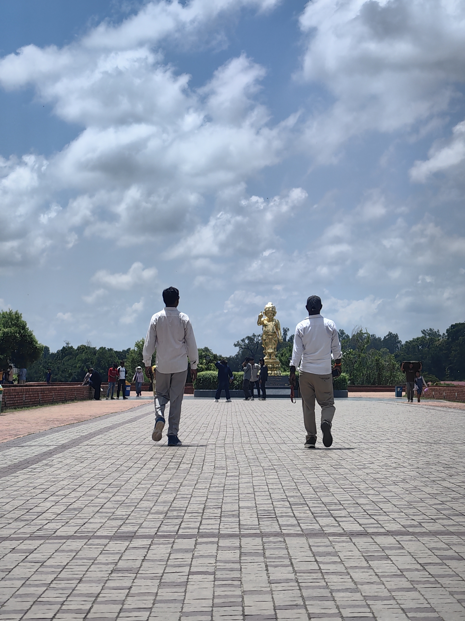 The image depicts an outdoor scene in Lumbini Bazaar, Nepal, under a partly cloudy sky. A wide paved walkway, laid with rectangular stone tiles, dominates the foreground, with two men walking away from the camera towards a golden statue of a child. Both men are wearing light-colored shirts and trousers. The man on the left is wearing a white shirt and gray trousers, while the man on the right has on a white shirt, brown trousers, and a black baseball cap. In the mid-ground, a golden statue of a child stands on a pedestal. Several other individuals are scattered across the walkway and the surrounding area, some walking, some standing. To the left, a red brick wall runs along the edge of the walkway, with lush green trees beyond. The background is filled with more trees and foliage. The overall impression is that of a public space, likely a tourist attraction, given the presence of the statue and the number of people.