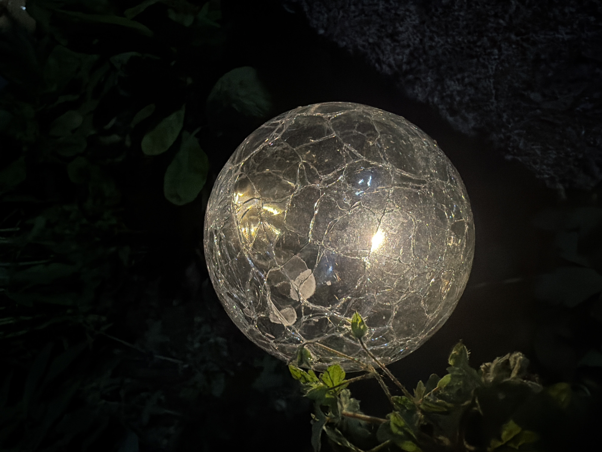 The image displays an outdoor nighttime scene, predominantly dark, featuring a centrally positioned, spherical illumination. The sphere is constructed from a transparent material with a pronounced "crackle glass" or "ice crack" texture, which disperses light internally. It emits a warm, yellowish-white glow, with two distinct brighter light sources visible within its core.
To the immediate left and partially in the foreground, vibrant green foliage from a plant is present, with some leaves and stems subtly illuminated by the sphere's light, revealing their texture and venation. The leaves appear slightly fuzzy in some areas. To the right and below the sphere, a dark, granular surface consistent with gravel or rough earth is visible. The background and upper portions of the image are enveloped in deep shadow, revealing only indistinct silhouettes of dark, leafy vegetation, suggesting a garden or natural setting. No individuals, animals, or discernable human activity are present. The image itself provides no discernible features or contextual information to confirm its alleged location in the city of Svetovrachene, Bulgaria.